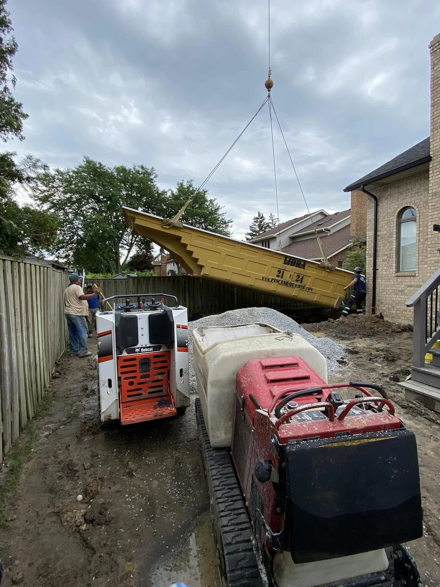 Construction workers are using a crane to lift a yellow boat into a backyard next to a brick house, with equipment like a skid steer loader and a mini excavator on a dirt surface.