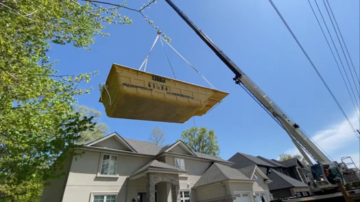 A fibreglass pool being lifted by a crane over residential houses with trees and power lines in the background.