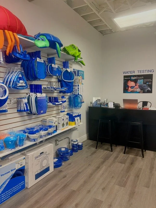 Scene inside a retail store or pool supply shop displaying various blue swimming pool accessories, including flotation devices, pool brushes, and pool maintenance supplies on wall-mounted racks. Two colorful floaties with cartoon characters are on top. A black checkout counter with a laptop and promotional poster for water testing is in the background.
