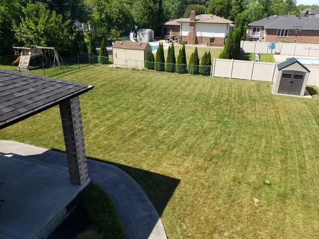 A before pool installation backyard with a well-maintained grass lawn, a small shed, and a fence with trees and neighboring houses in the background.