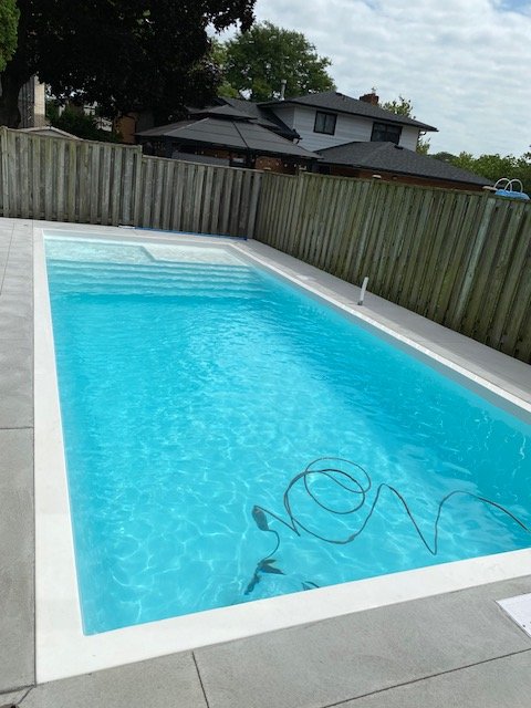 Empty backyard swimming pool with a cleaning tool and hose, fenced yard, neighboring house in the background.
