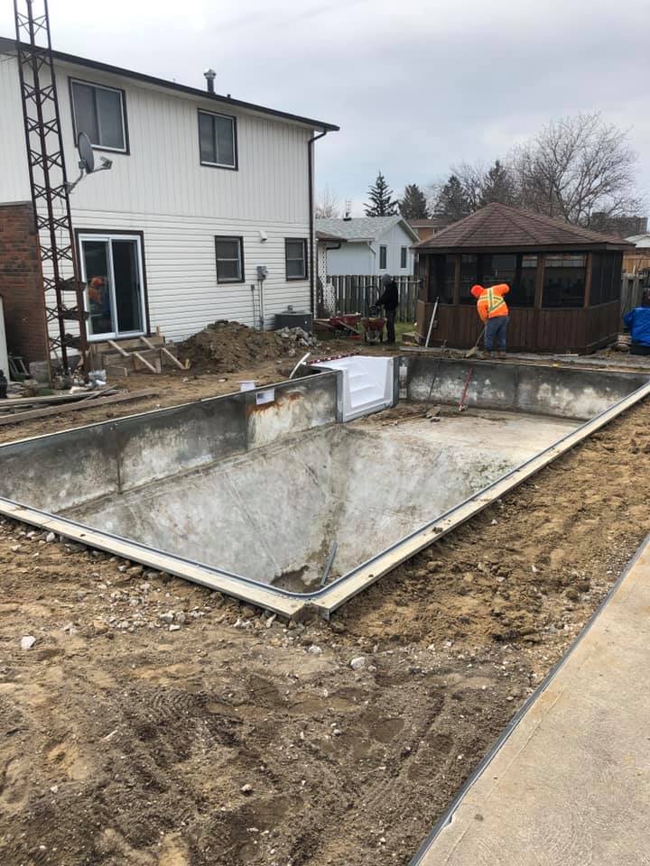 Pool renovation, showing and in-ground empty swimming pool under construction in backyard, with workers in safety vests, residential houses in background,