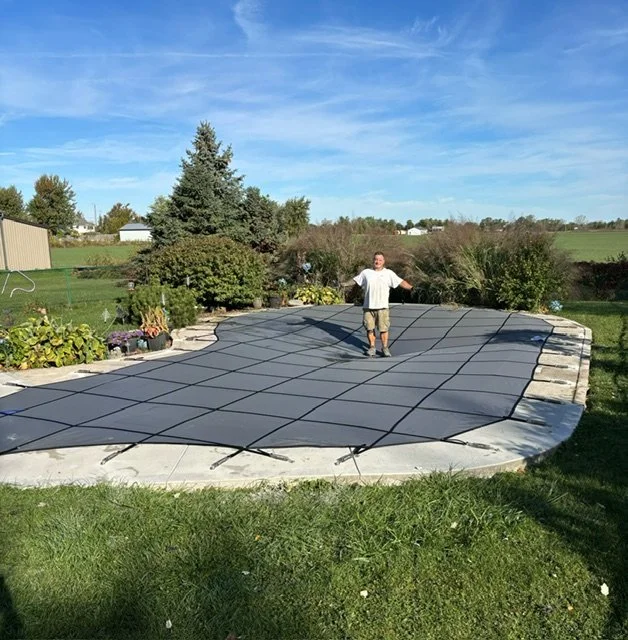 Person standing on a covered swimming pool in a backyard with trees and plants.