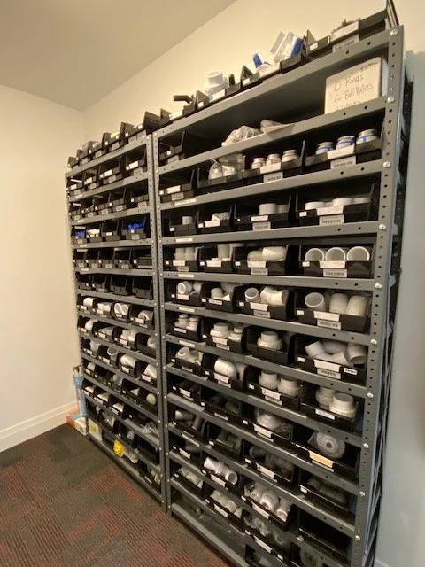 Shelves filled with various sizes and types of white PVC pipes and fittings, organized in black storage trays in a storage room.