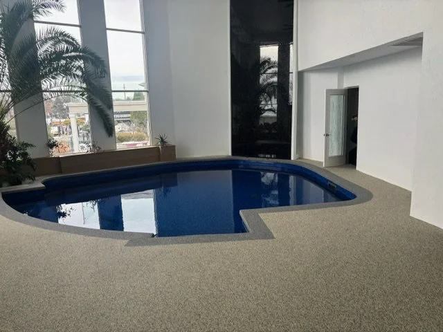 Indoor swimming pool with blue water, surrounded by beige pool deck, with large windows and some plants in the background.