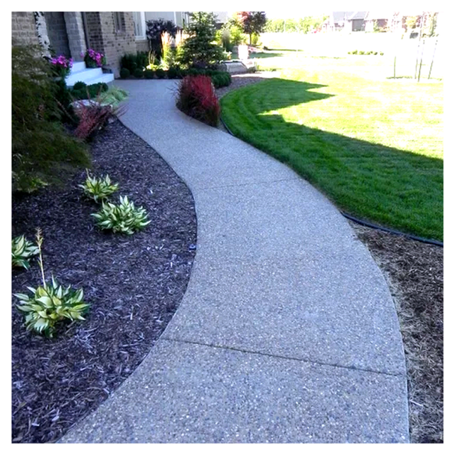 Curved exposed aggregate concrete walkway adjacent to a landscaped garden with various plants and a well-maintained lawn in a residential yard.