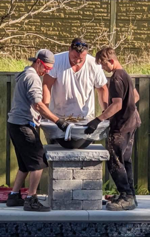 Two boys and an adult man working together on a firepit project outdoors, with a wooden fence and trees in the background.
