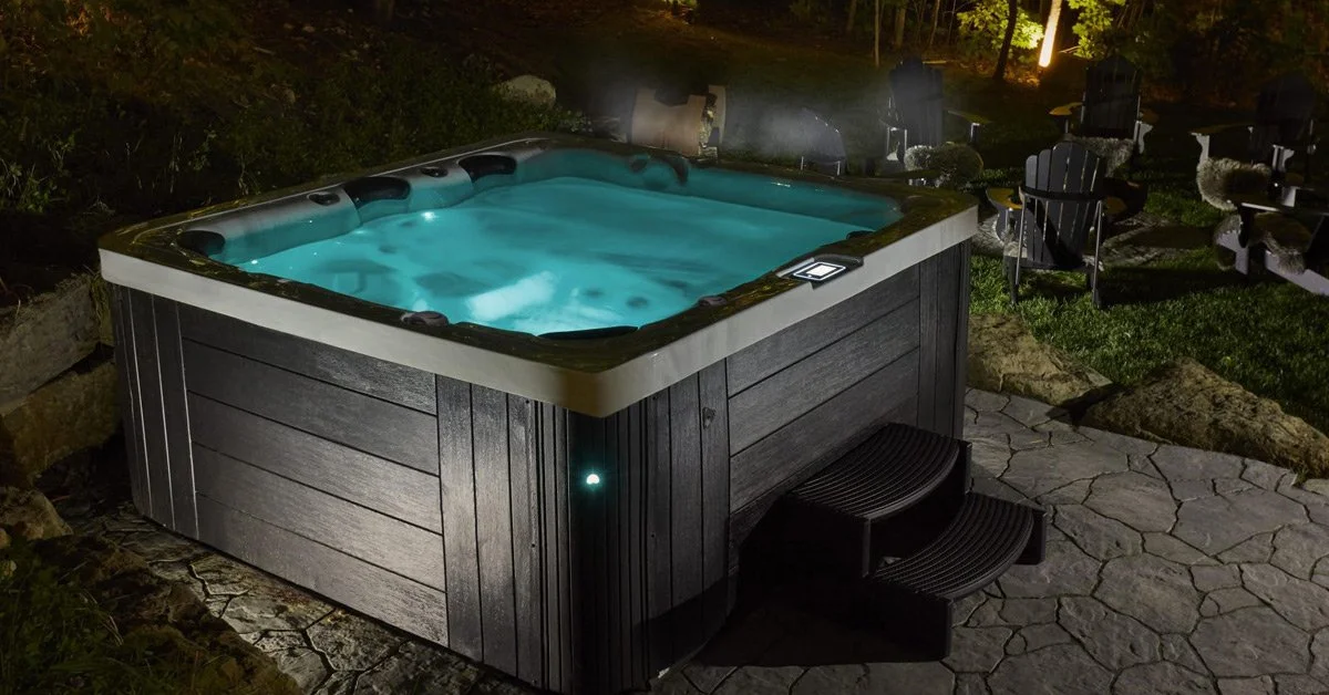 A hot tub with lit water on a stone patio at night, surrounded by chairs, rocks, and garden lights.