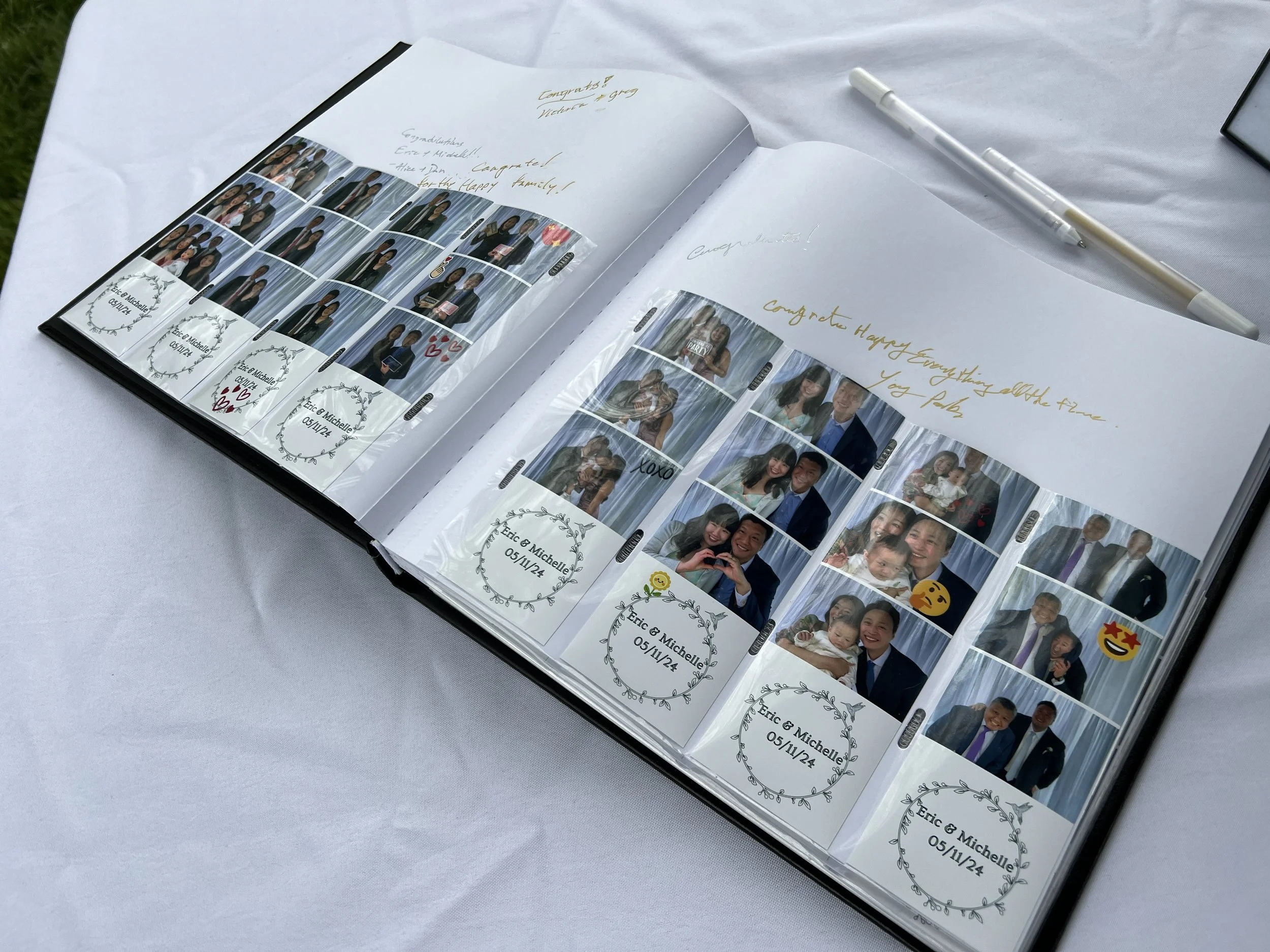 Open photo booth album with photo strips of people at a celebration, on a white table with a white pen, in an outdoor setting.