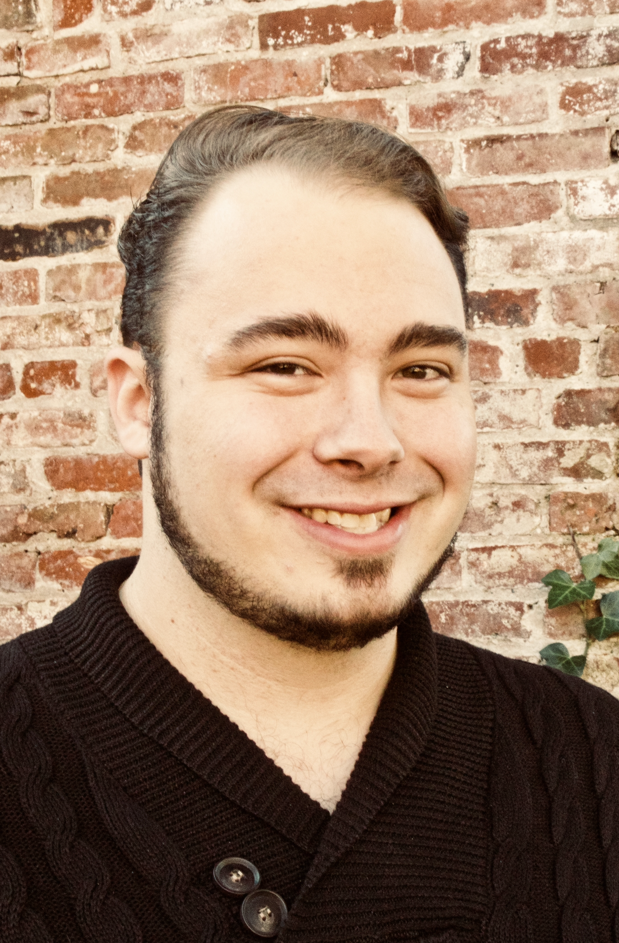 A smiling young man with dark hair and facial hair, wearing a black button-up sweater, standing in front of a brick wall with some green ivy.