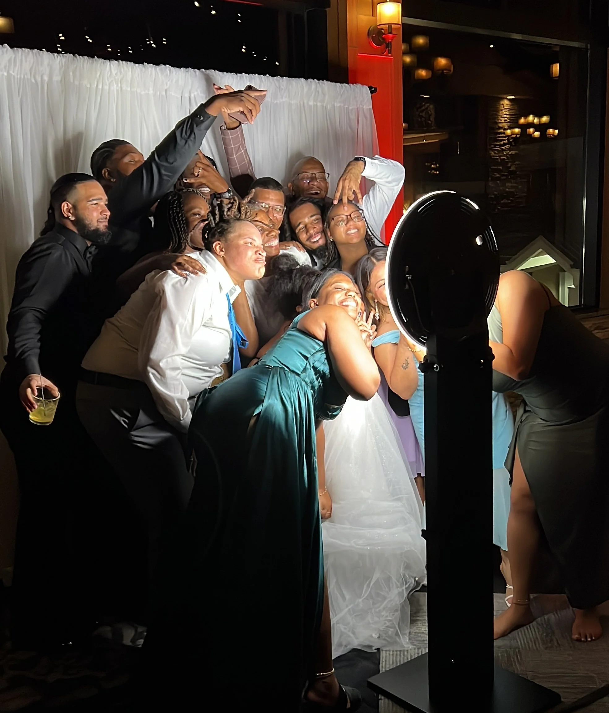 A group of people taking a group selfie with a photo booth at a wedding reception. They are smiling and appear to be happy.