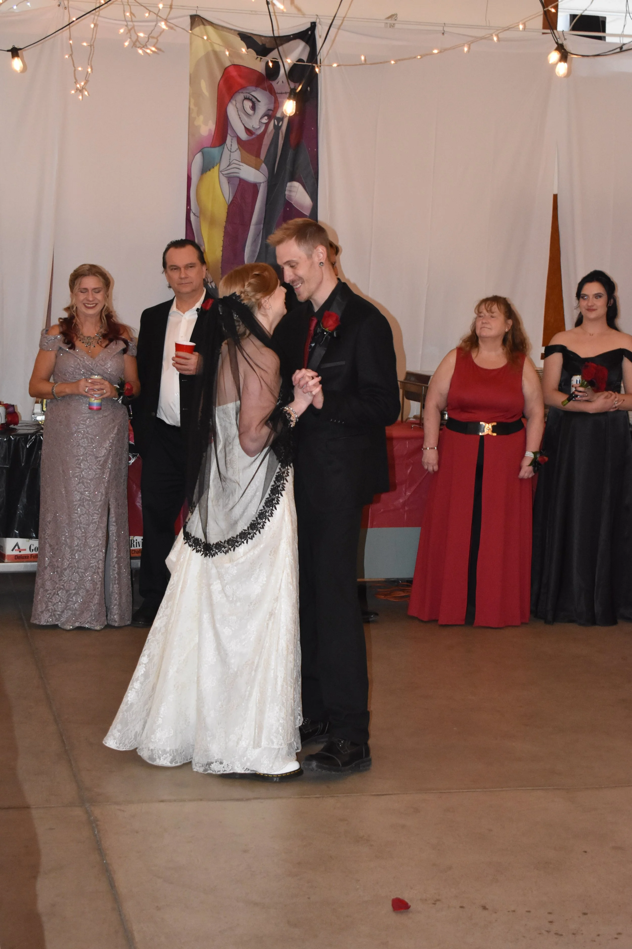 A wedding dance with a bride and groom holding hands, surrounded by five women in evening dresses, with a colorful Bride of Eternal Night poster in the background and string lights decorating the ceiling.