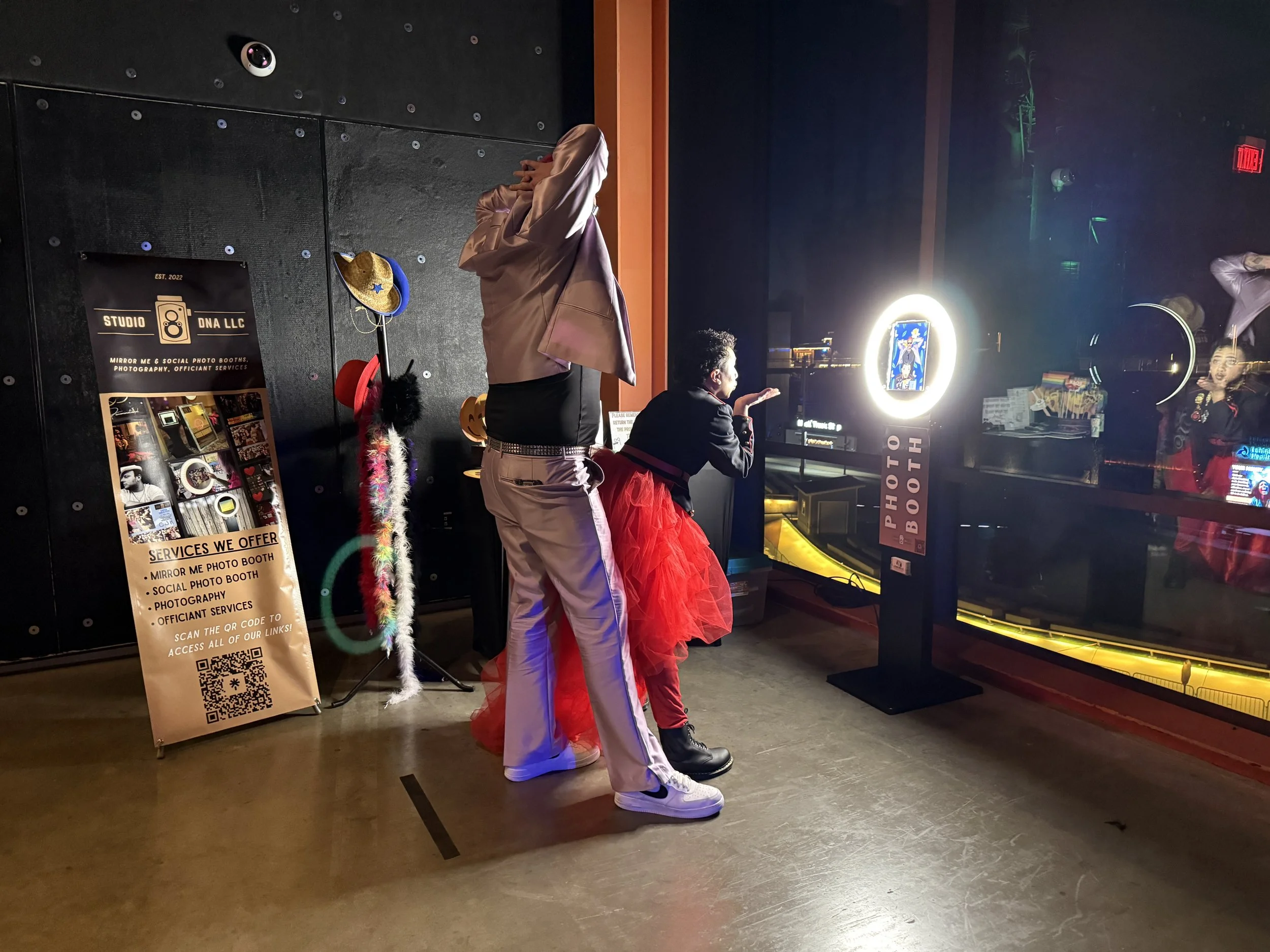 People in costume at a photo booth, with props like hats and feather boas, at a social event in a dimly lit room.