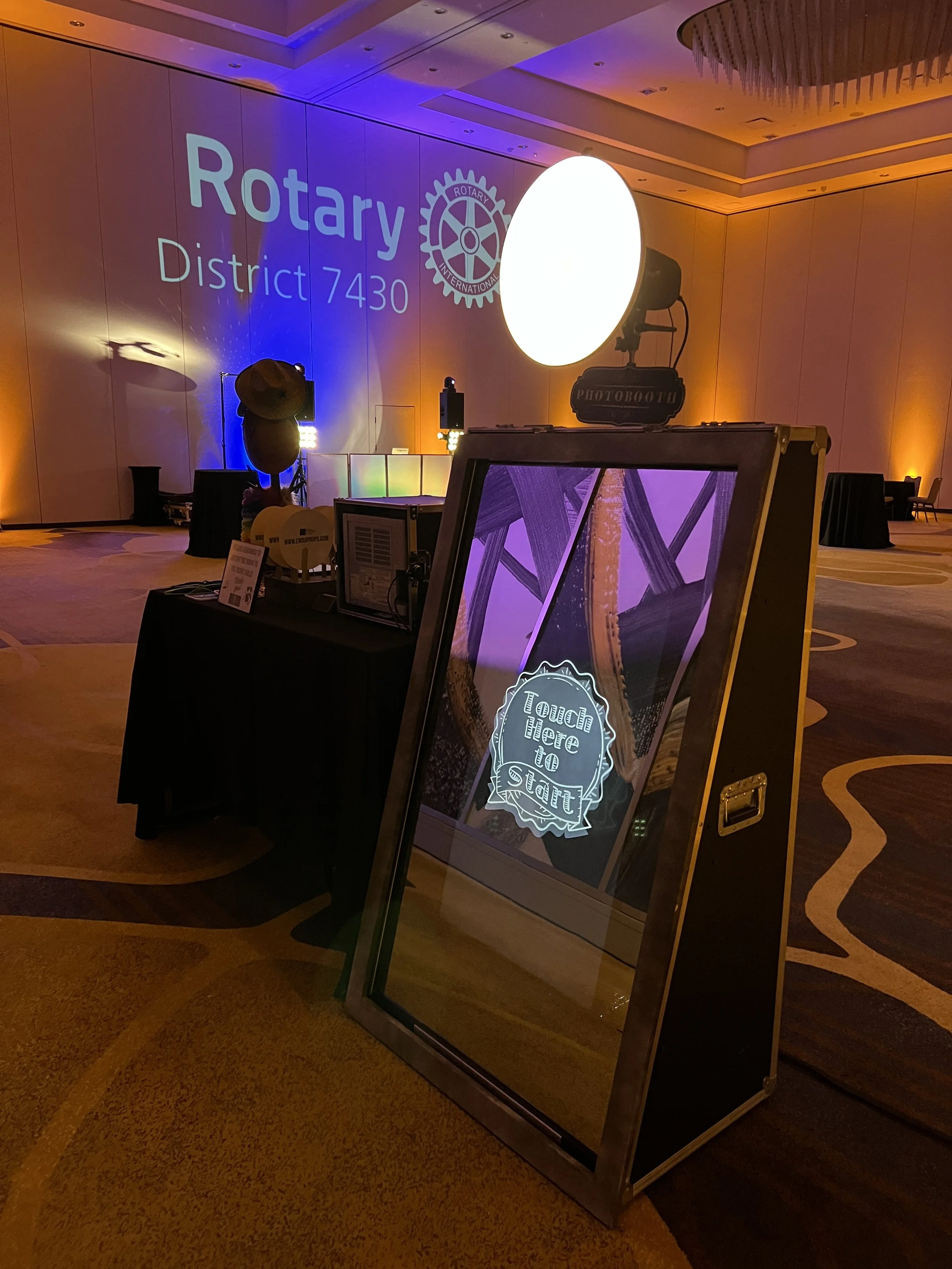 Event space with a large circular illuminated display, a sign reading 'Touch here to start,' and a decorative sign with a gear and 'Rotary District 7430' projected on a wall.
