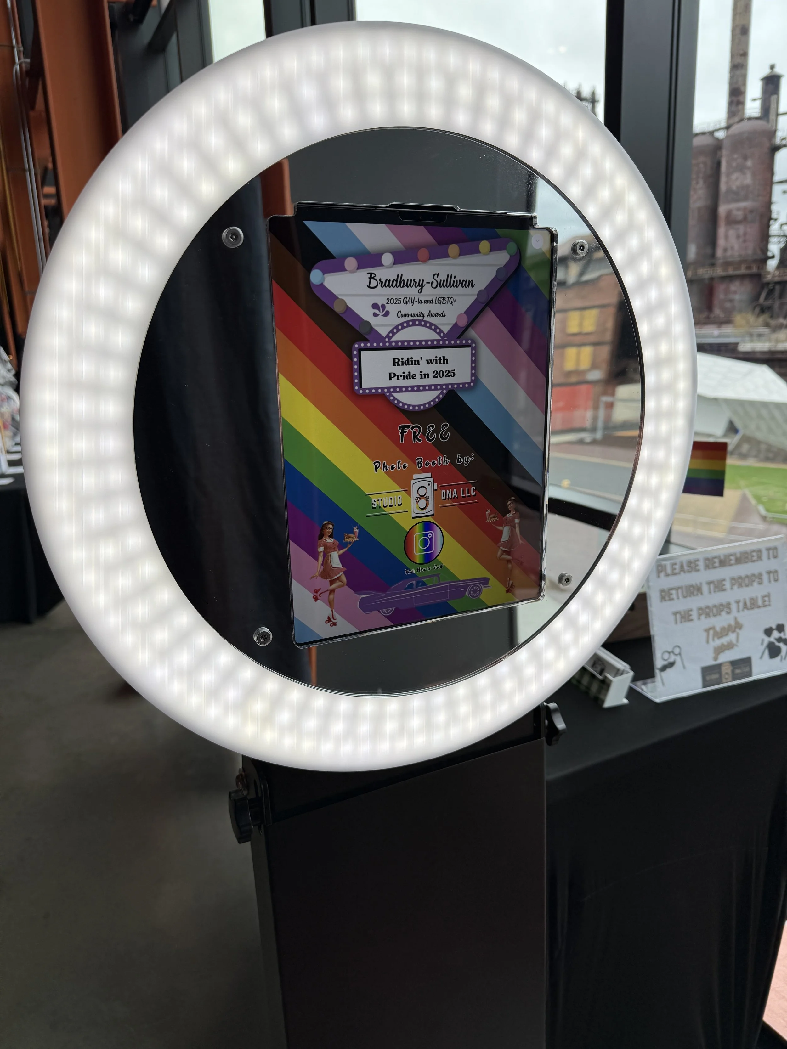 A photo booth with a ring light surrounding a smartphone displaying a colorful Pride-themed graphic for the 2025 Gay and LGBTQ+ Community Awards, with text reading 'Ridin' with Pride in 2025' and an illustration of a woman in a retro outfit standing by a vintage car.