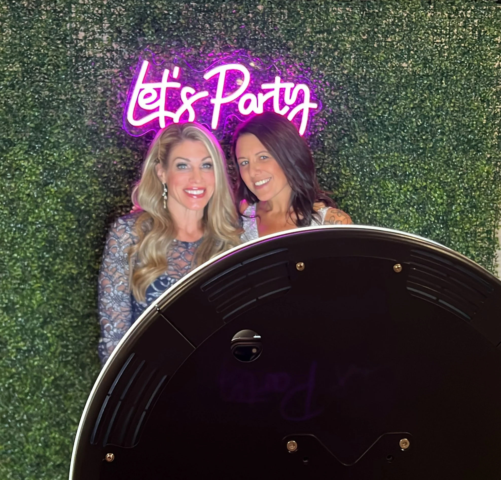 Two women taking a selfie in front of a green wall with a neon sign that reads 'Let's Party' in purple and pink colors, with part of a black photo booth visible in the foreground.