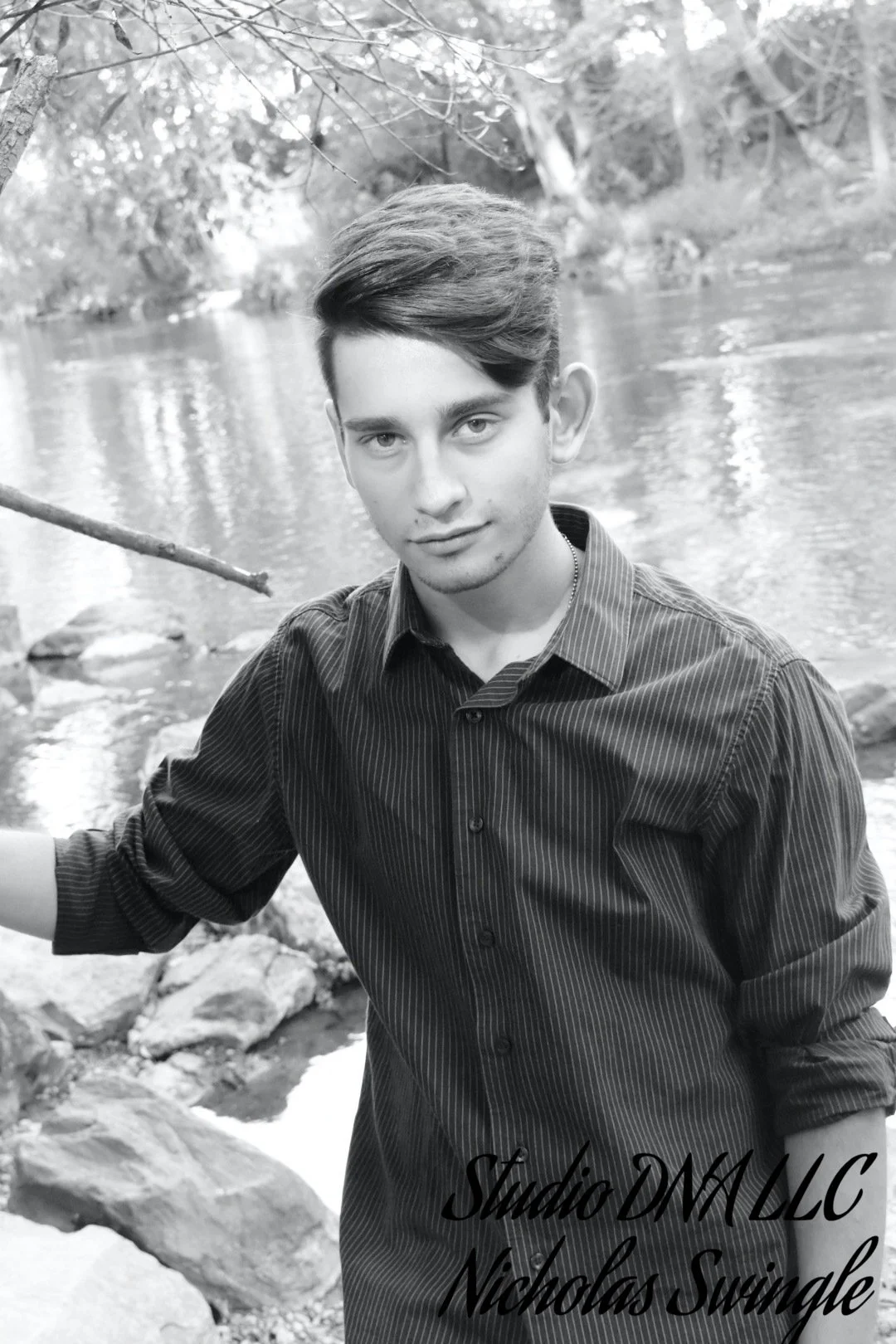 A young man with styled hair and a striped shirt standing outdoors near a river, with rocks and trees in the background, black-and-white photo.