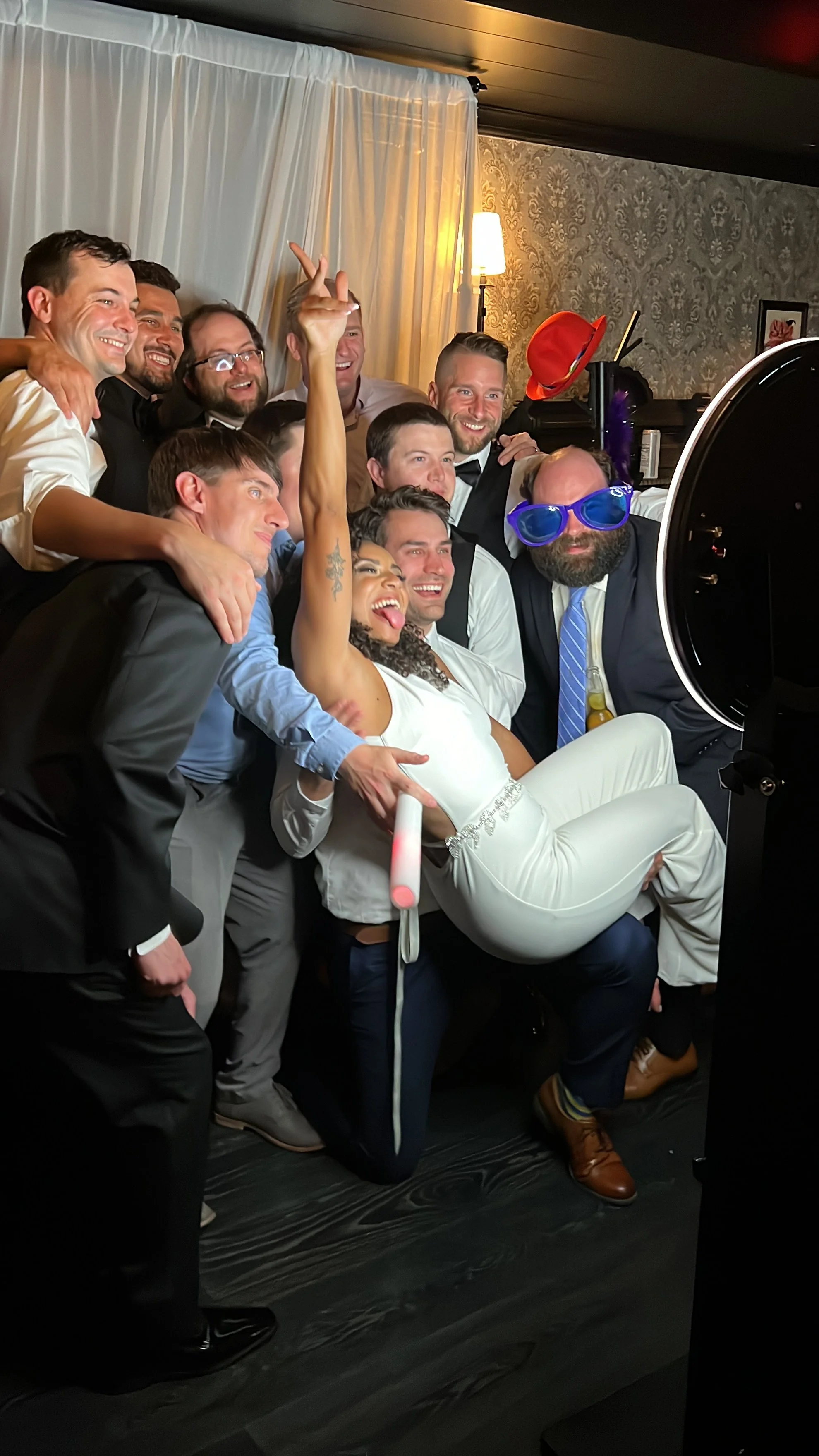 A group of friends taking a photo together at a celebration or party. They are smiling, posing, and some are wearing fun accessories like oversized glasses and a red hat.