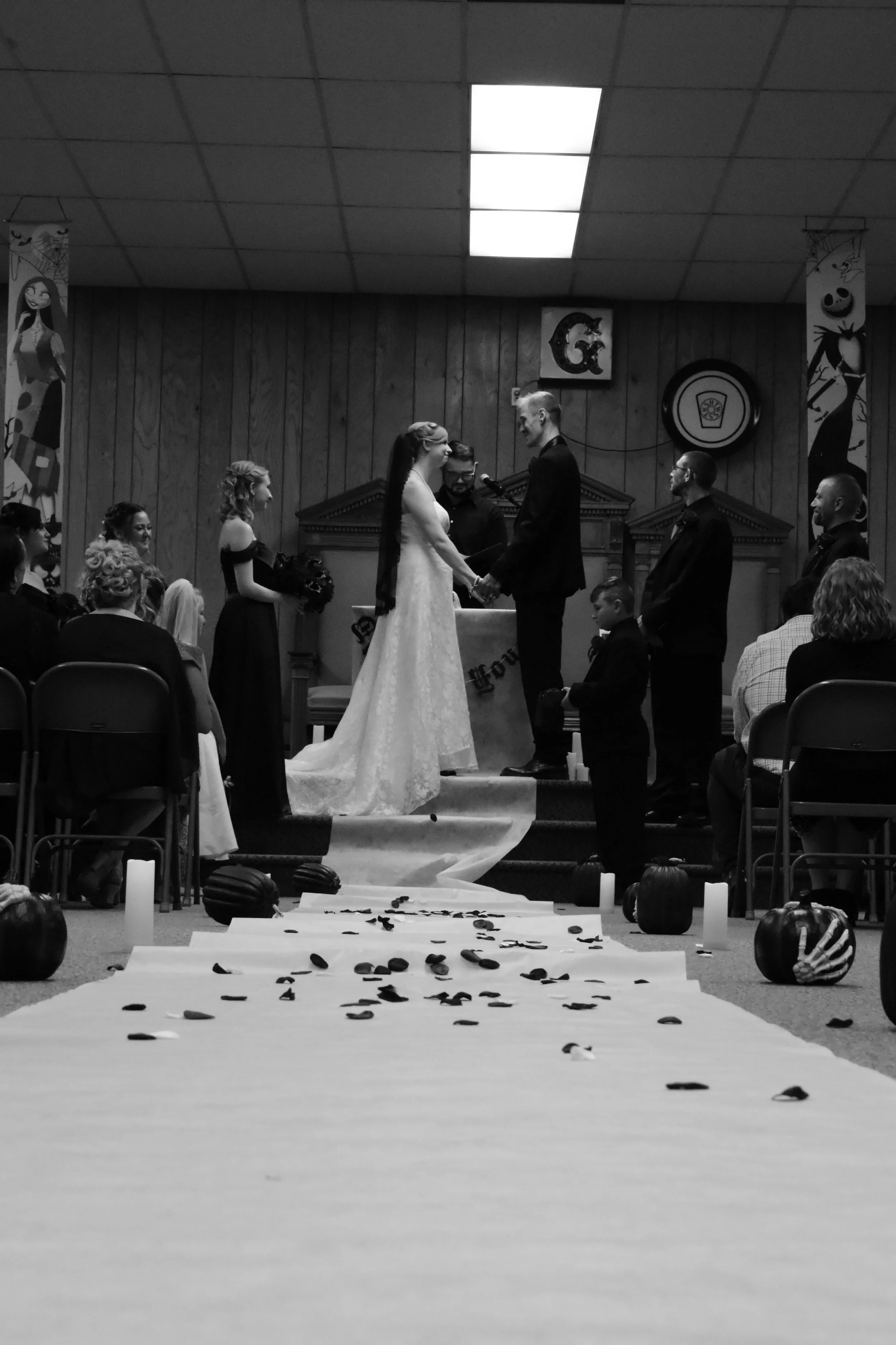 A black and white photo of a wedding ceremony with a bride and groom holding hands at the altar, surrounded by bridesmaids and groomsmen. The aisle is decorated with pumpkins, candles, and scattered black petals. An officiant is in the background.