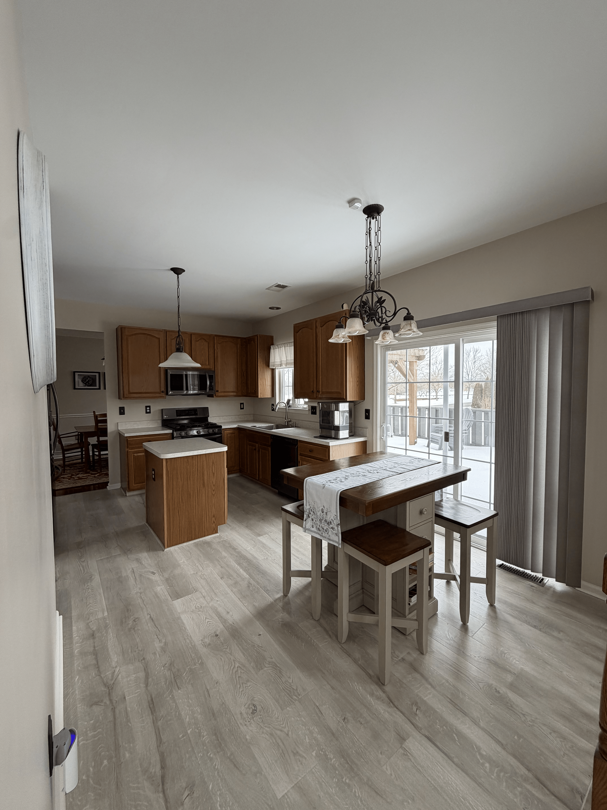 A kitchen with wooden cabinets, a dining table with a lace table runner, white chairs, and sliding glass doors leading to a snowy outdoor scene.