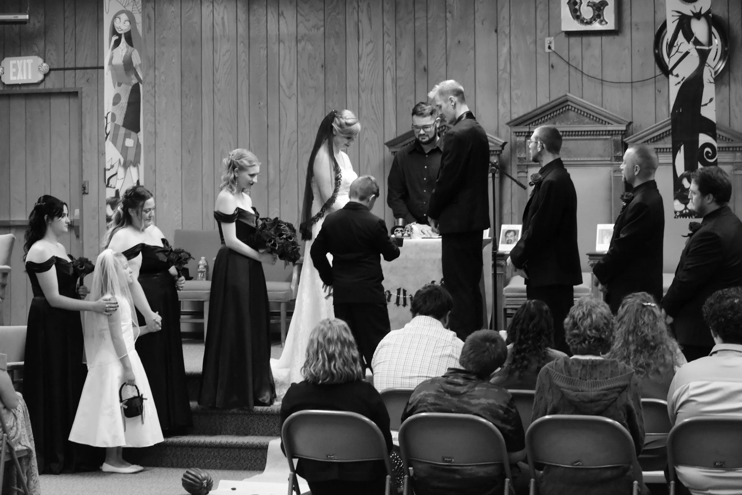 People participating in a wedding ceremony inside a wooden-paneled hall. The bride and groom are standing in front of officiants, with bridesmaids and groomsmen standing beside them. The scene is formal with the bride in a white gown and the groom and groomsmen in dark suits wearing boutonnières. Several guests are seated, observing the ceremony.