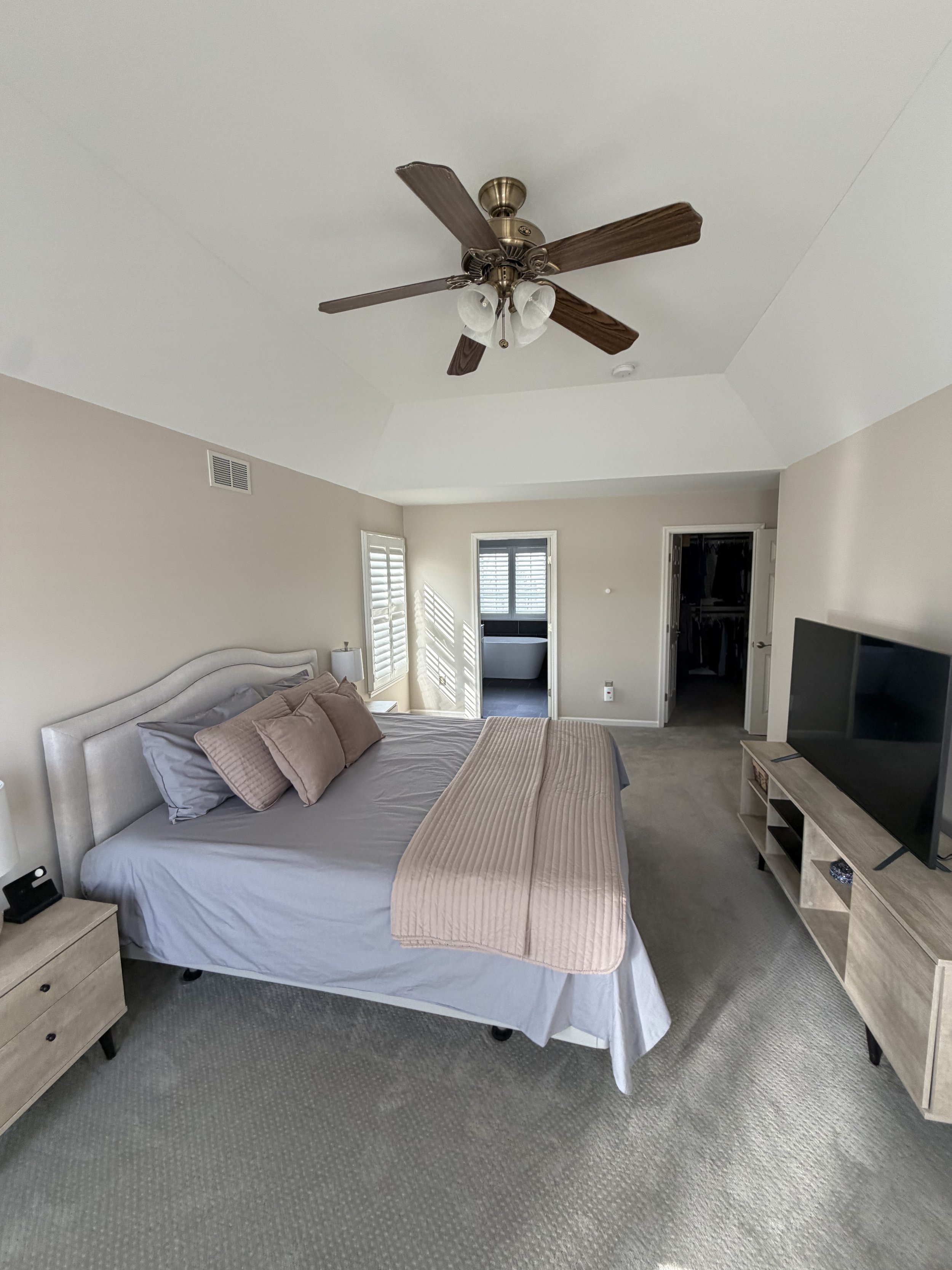 A bedroom with a bed, nightstand, and television, with ceiling fan and windows with shutters.