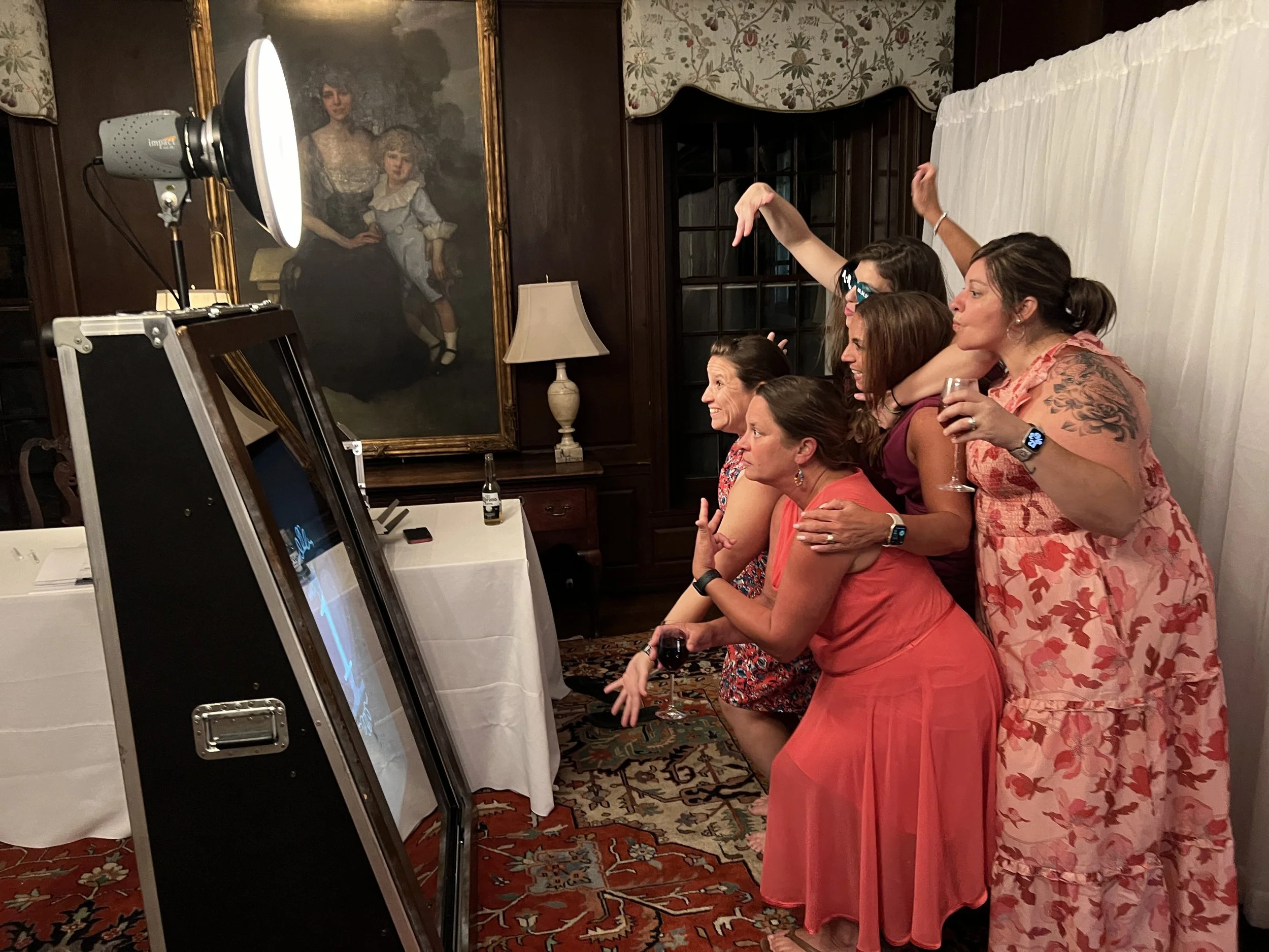 A group of women at a party playing a photo booth game with a vintage portrait painting in the background.