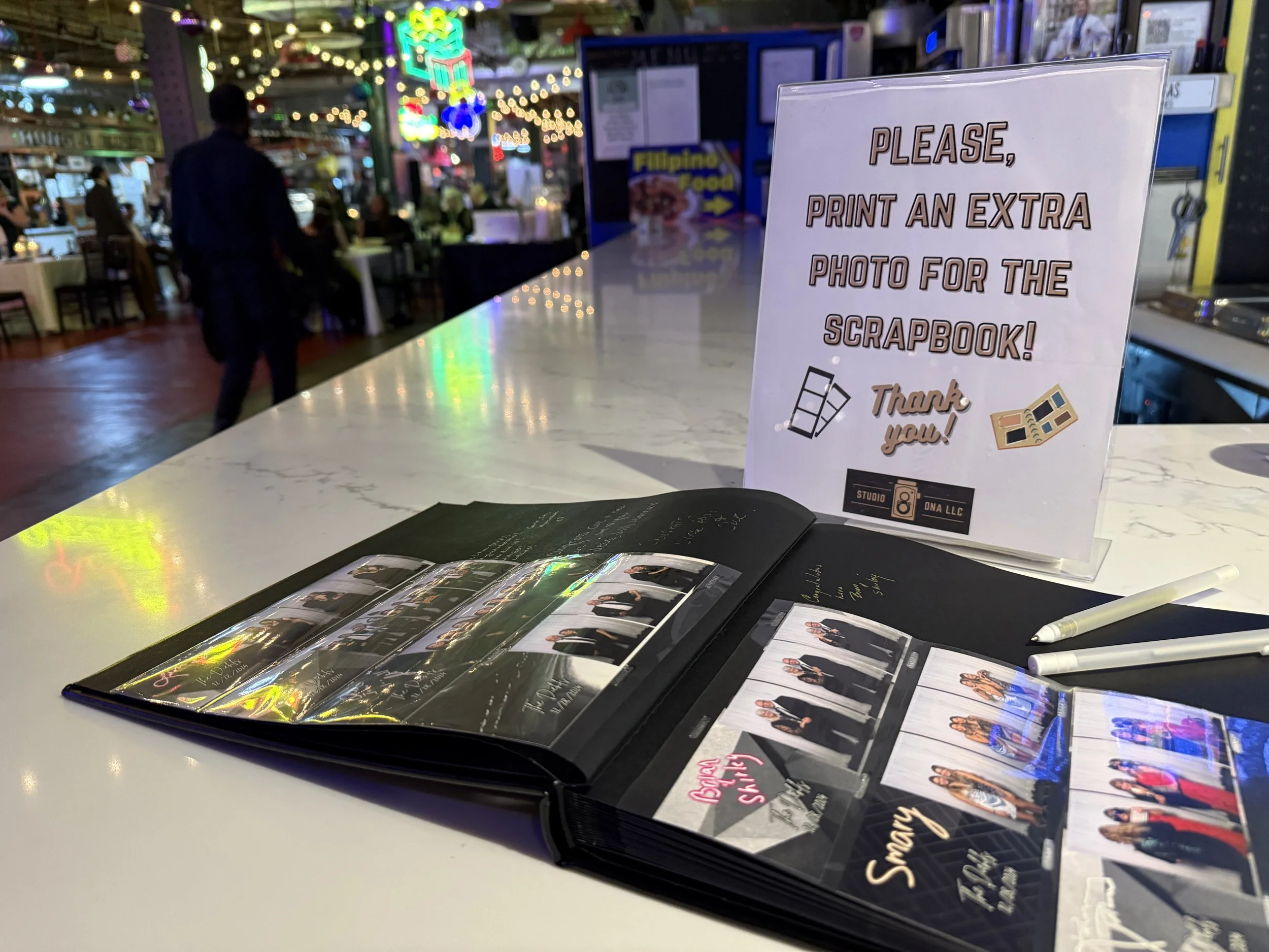 A white table with photo books and pictures of a woman on a black background, with a sign that says, 'Please, print an extra photo for the scrapbook!' in a busy, indoor location with people and colorful lights in the background.