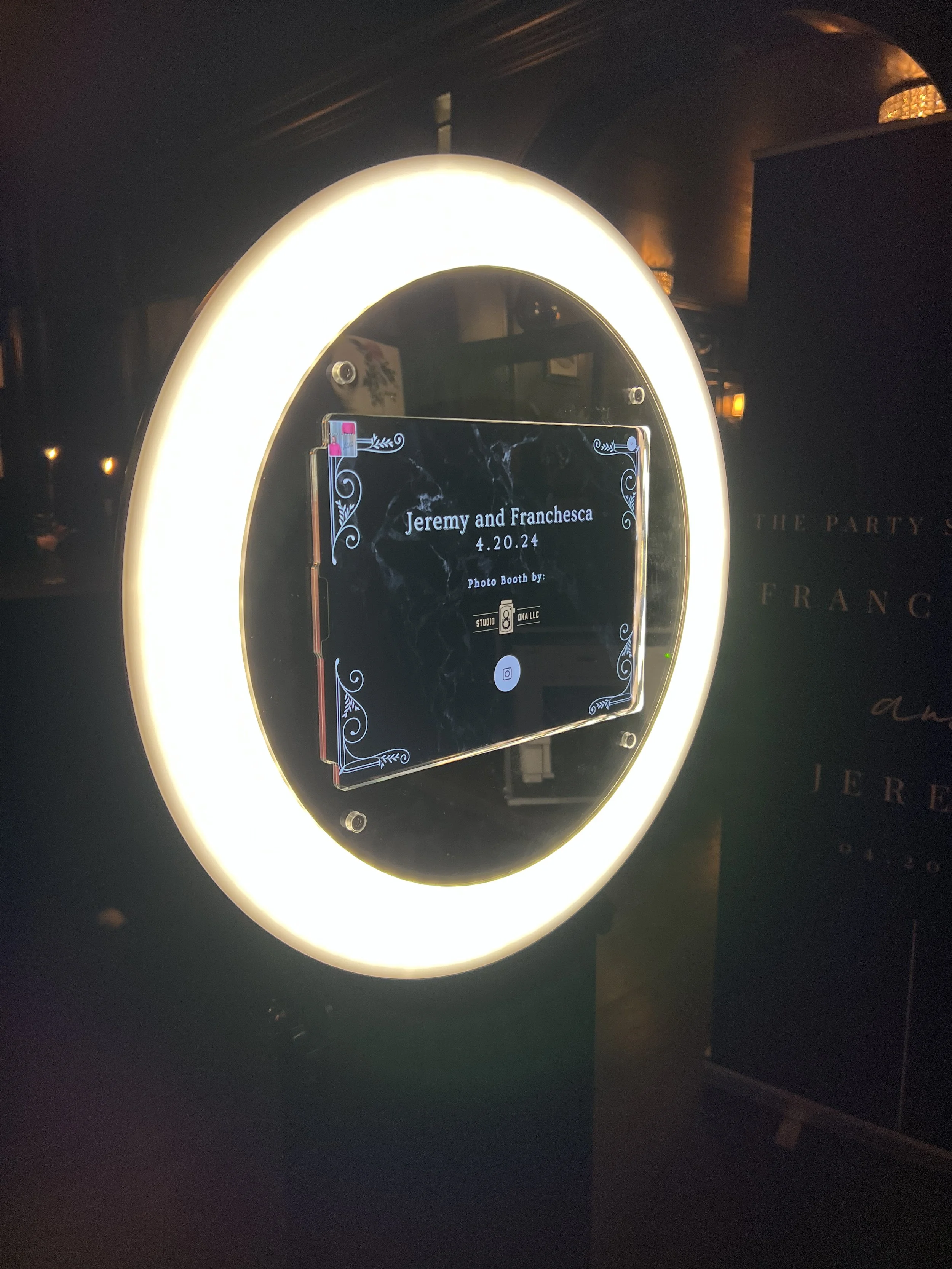 Photo booth with illuminated ring border, display screen showing event details for Jeremy and Franchesca wedding on April 20, 2024, in a dimly lit setting.