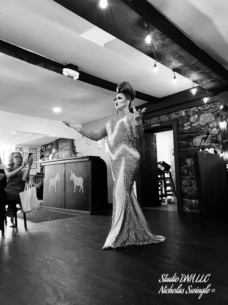 A drag queen in a sparkling, form-fitting gown performing on a stage in a bar or club, with an audience member taking a photo.