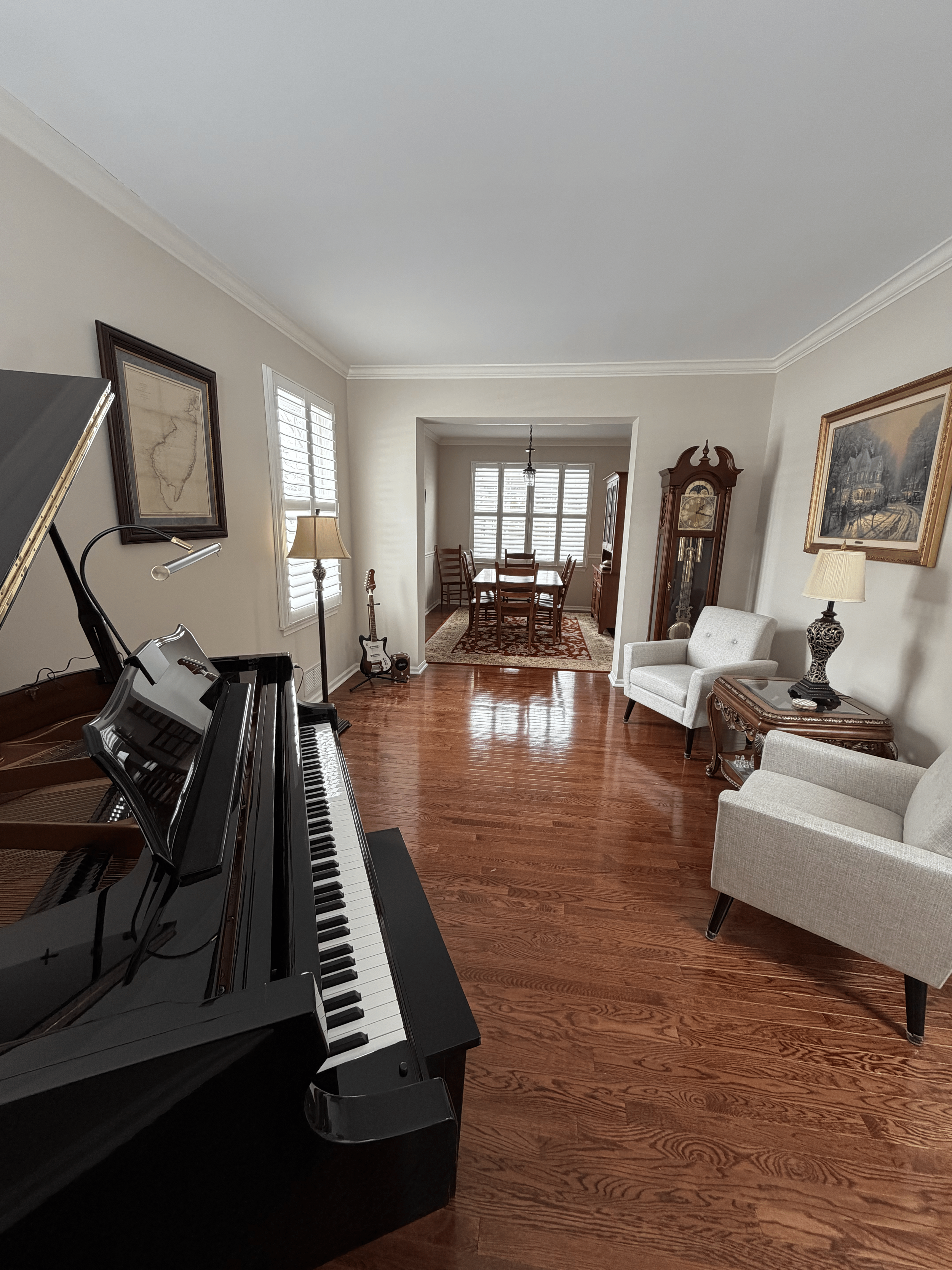 Living room with a black grand piano in the foreground, white armchair to the right, two table lamps, a wooden side table, antique wall clock, a framed map on the wall, guitar near window with shutters, and artwork of a house in winter on the wall.