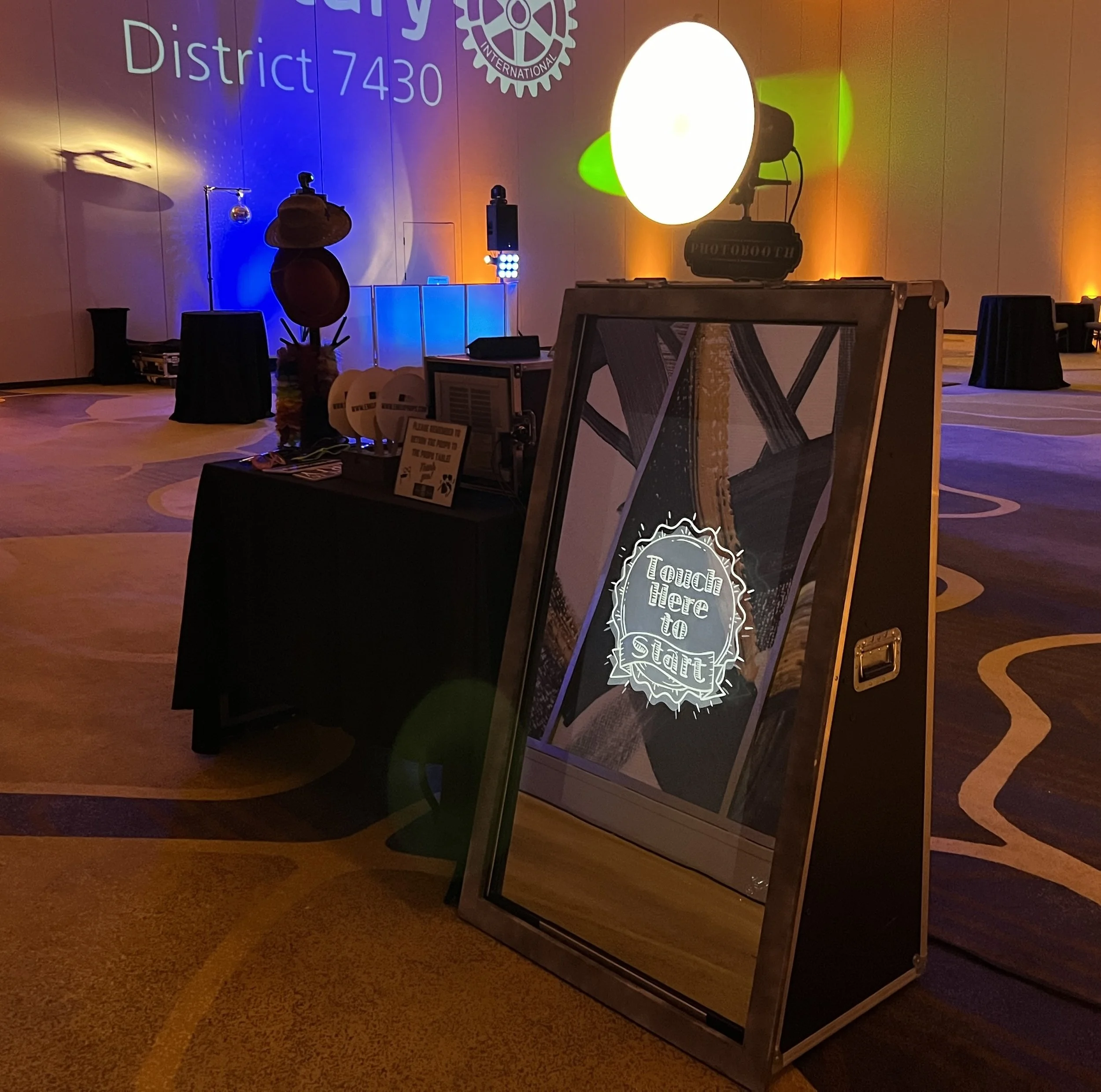 Event setup with a large illuminated sign that reads 'Touch here to start,' a wooden table, various equipment, and a backdrop displaying the Rotary International logo and the text 'District 7430.' The space has warm lighting with purple, blue, yellow, and orange hues.