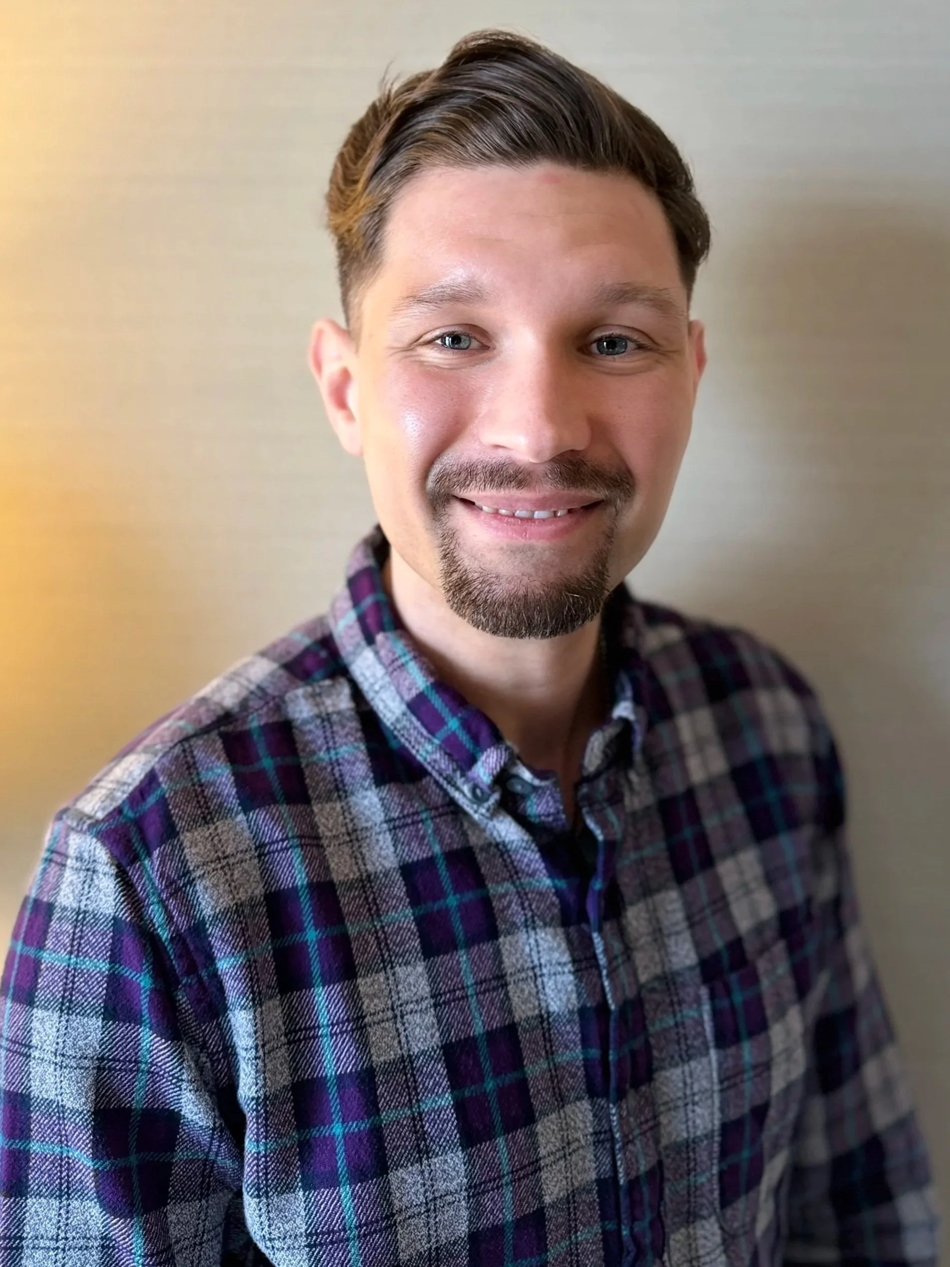 A young man with light skin, short brown hair, blue eyes, and facial hair, smiling while wearing a plaid shirt.