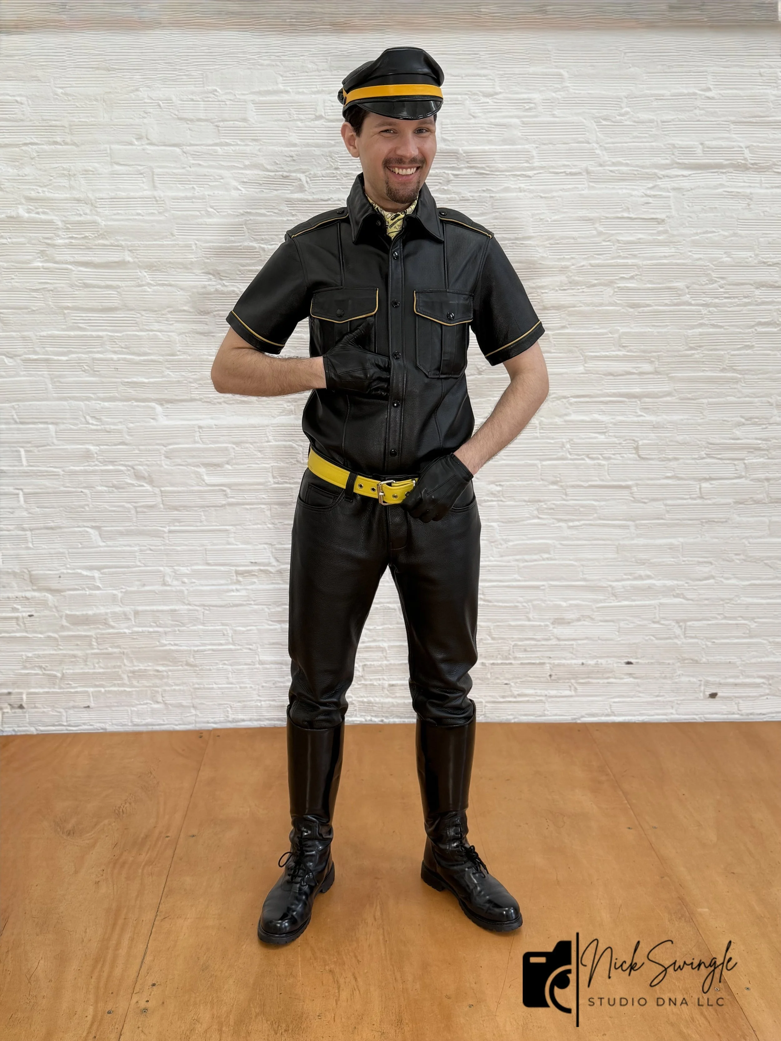 A man smiling dressed in a black leather formal outfit with yellow accents, black gloves, black pants, and black boots, standing in front of a textured white wall and wooden floor.