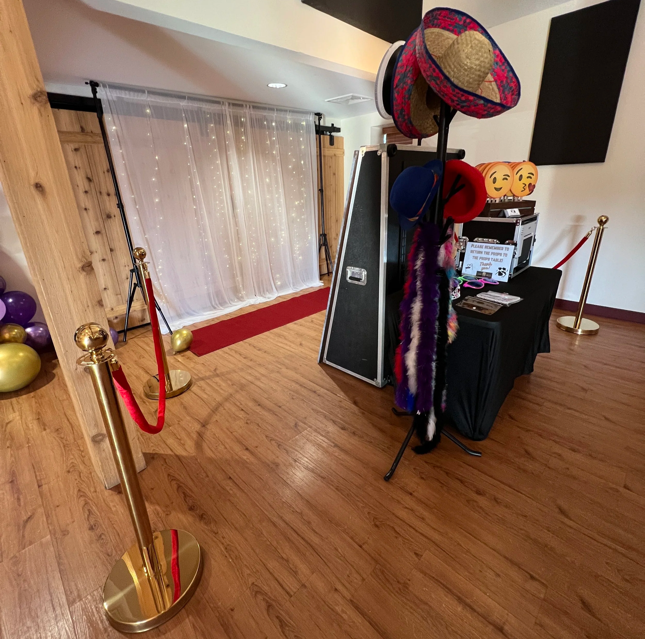 Photo of a photo booth setup with a red carpet, a backdrop of fairy lights and balloons on the floor, a table with props including colorful hats and emoji masks, and a sign on the table with instructions for guests.