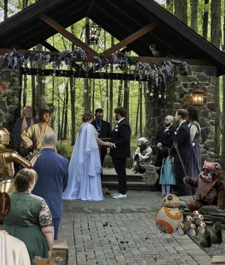 A wedding ceremony taking place outdoors in a forest setting under a wooden pavilion with stone columns, decorated with purple flowers and hanging star-shaped ornaments. The bride and groom stand front and center, holding hands, with guests and characters from Star Wars, including a robot and a teddy bear, observing.
