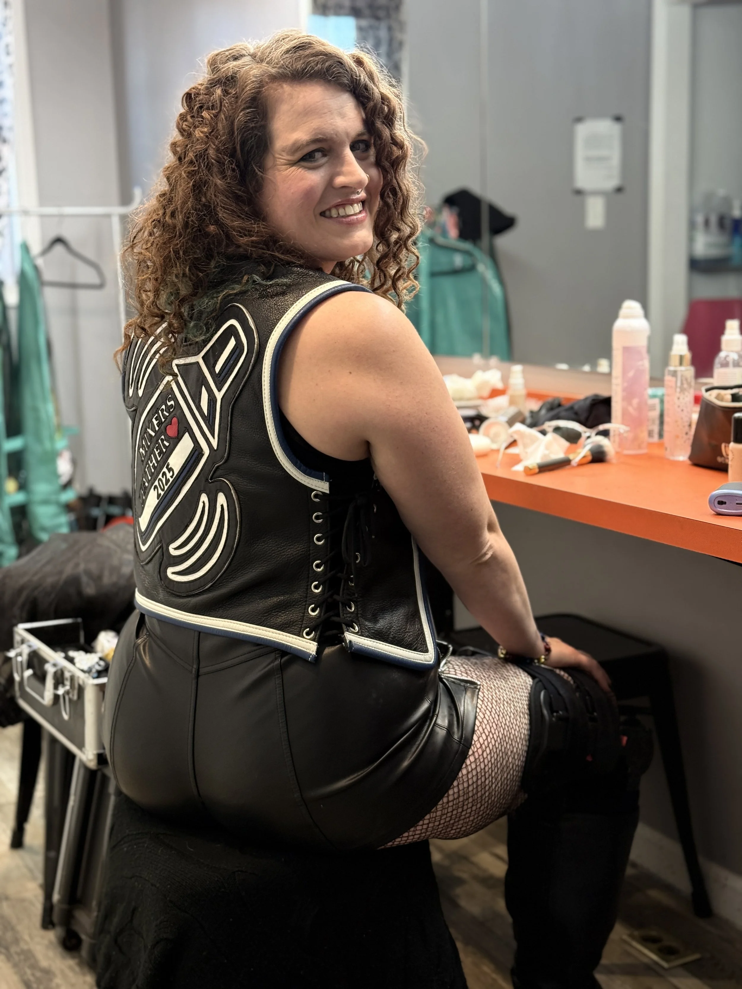 A woman with curly hair smiling and looking at the camera, wearing a black motorcycle vest and black leather skirt, sitting in a dressing room or backstage area with makeup and beauty products on the table nearby.