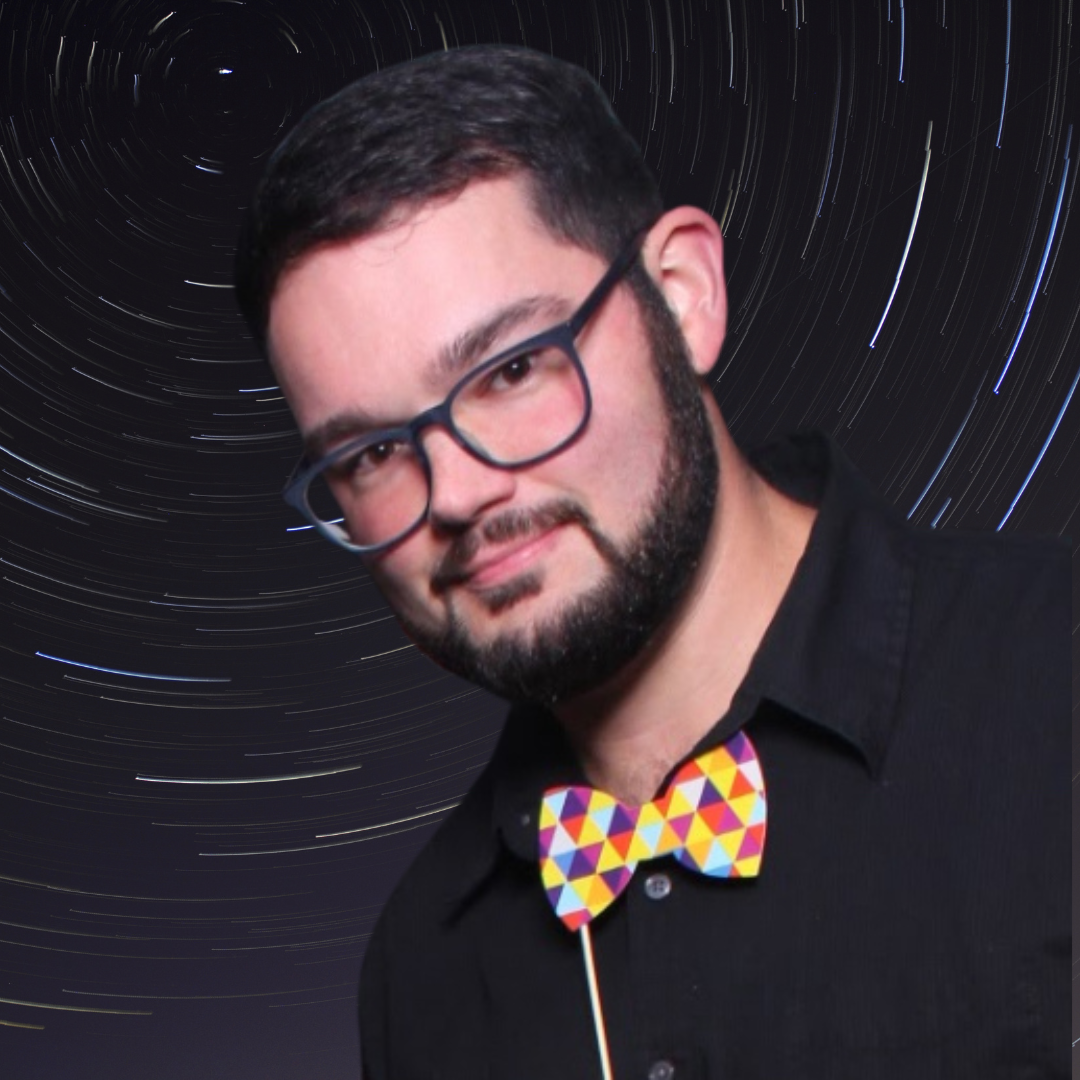 A man with glasses, a beard, and mustache wearing a black shirt and colorful geometric bow tie, standing in front of a star trail night sky background.
