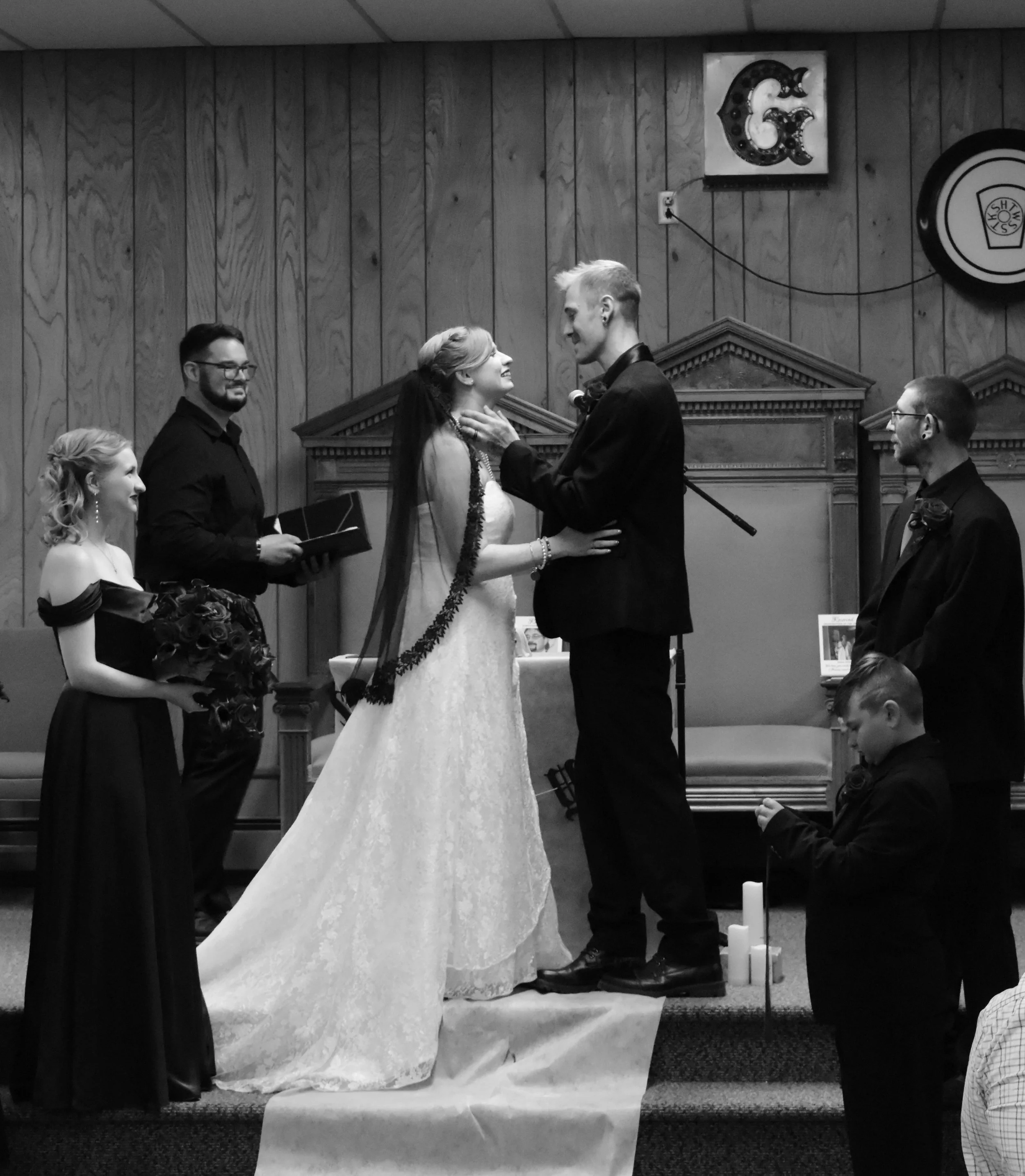 A wedding ceremony with a bride and groom holding hands and gazing at each other, surrounded by attendants, an officiant, candles, and decorative elements in a wood-paneled room.