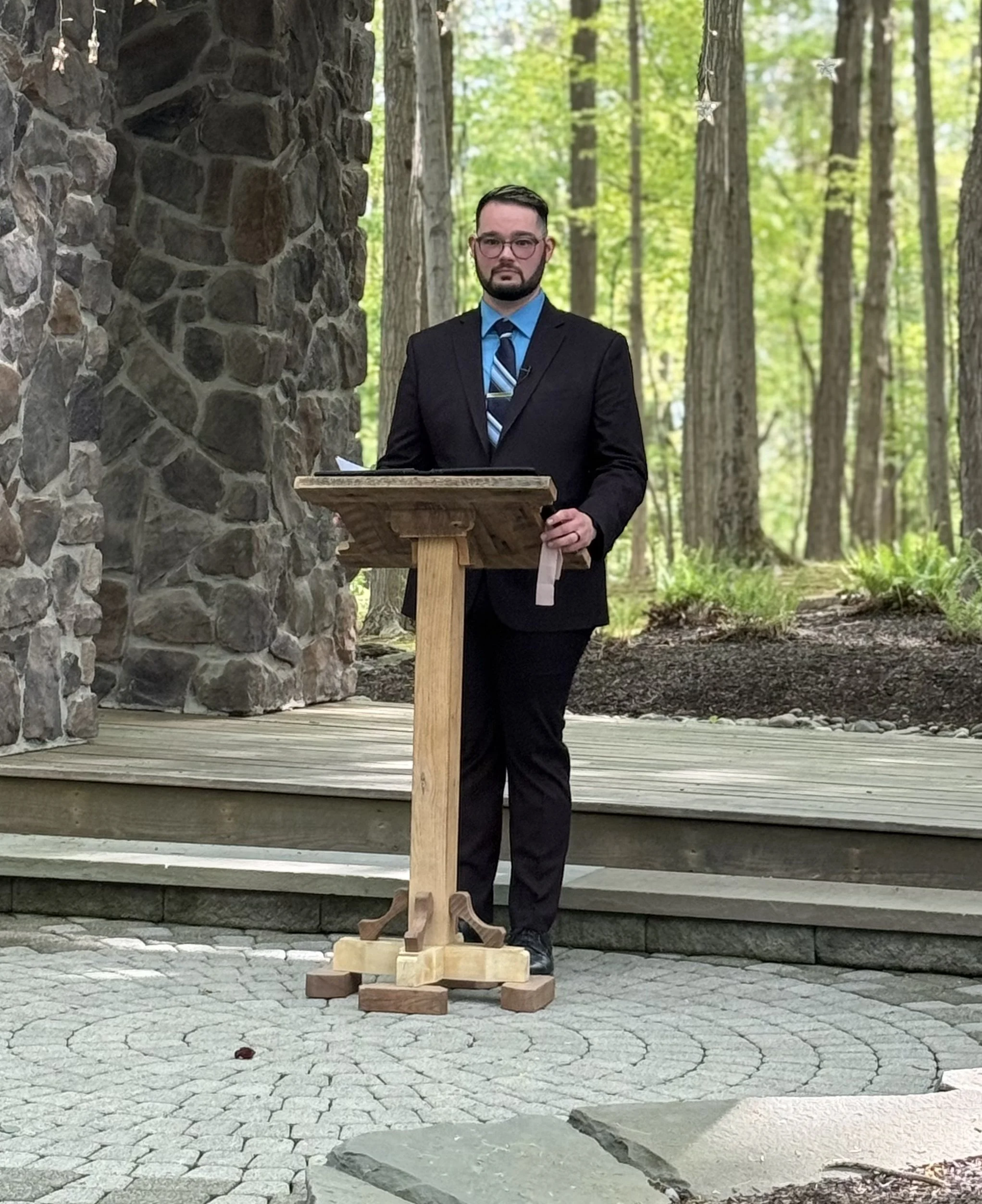 A man in a black suit, blue shirt, and striped tie stands behind a wooden podium outdoors in a forest with green trees. A stone structure and star-shaped decorations are visible nearby. He is about to officiate a wedding.