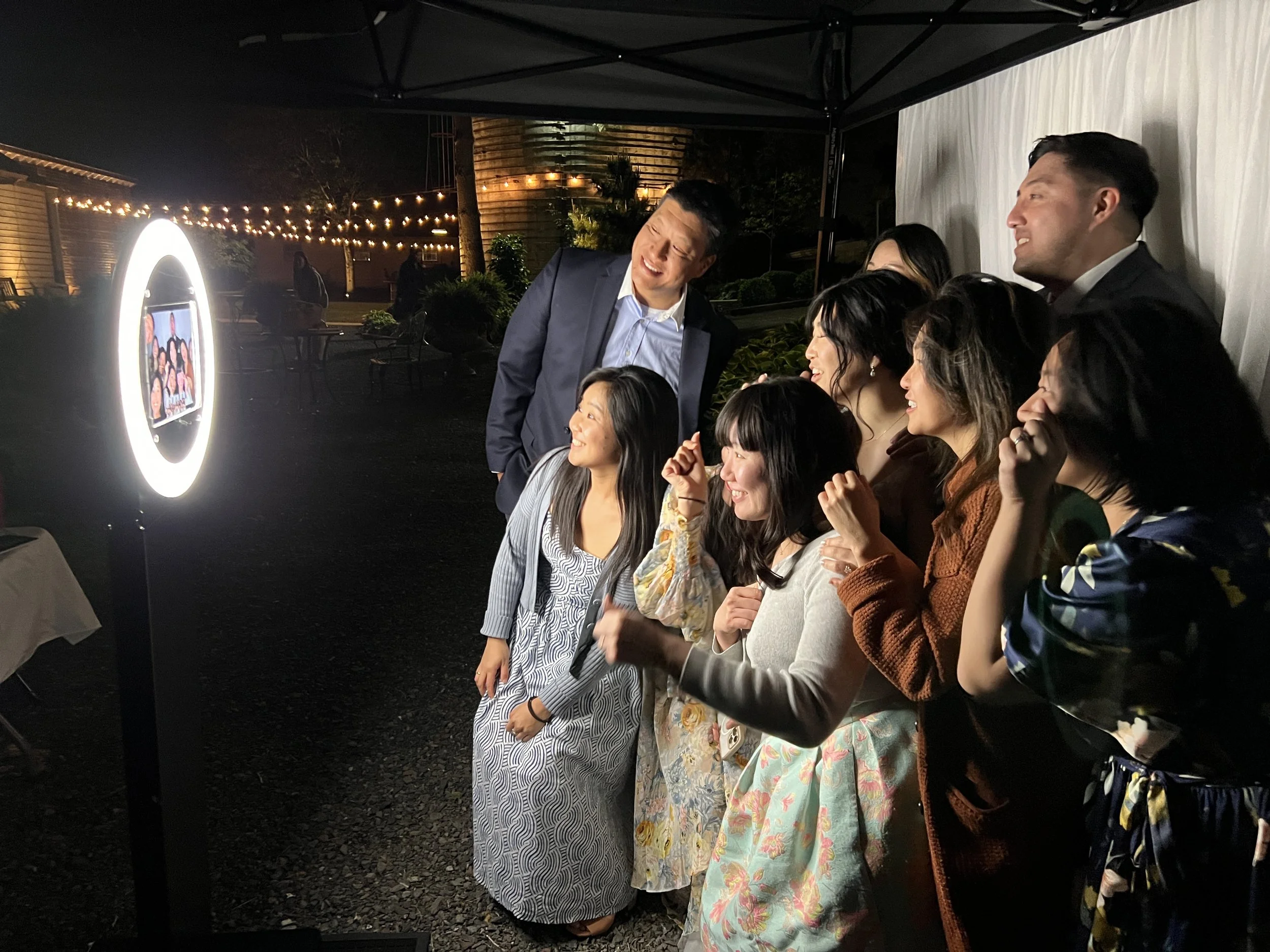 Group of people gathered at a photo booth during an outdoor evening event, smiling and posing for a picture. A photo frame with an illuminated border is visible on the left side.
