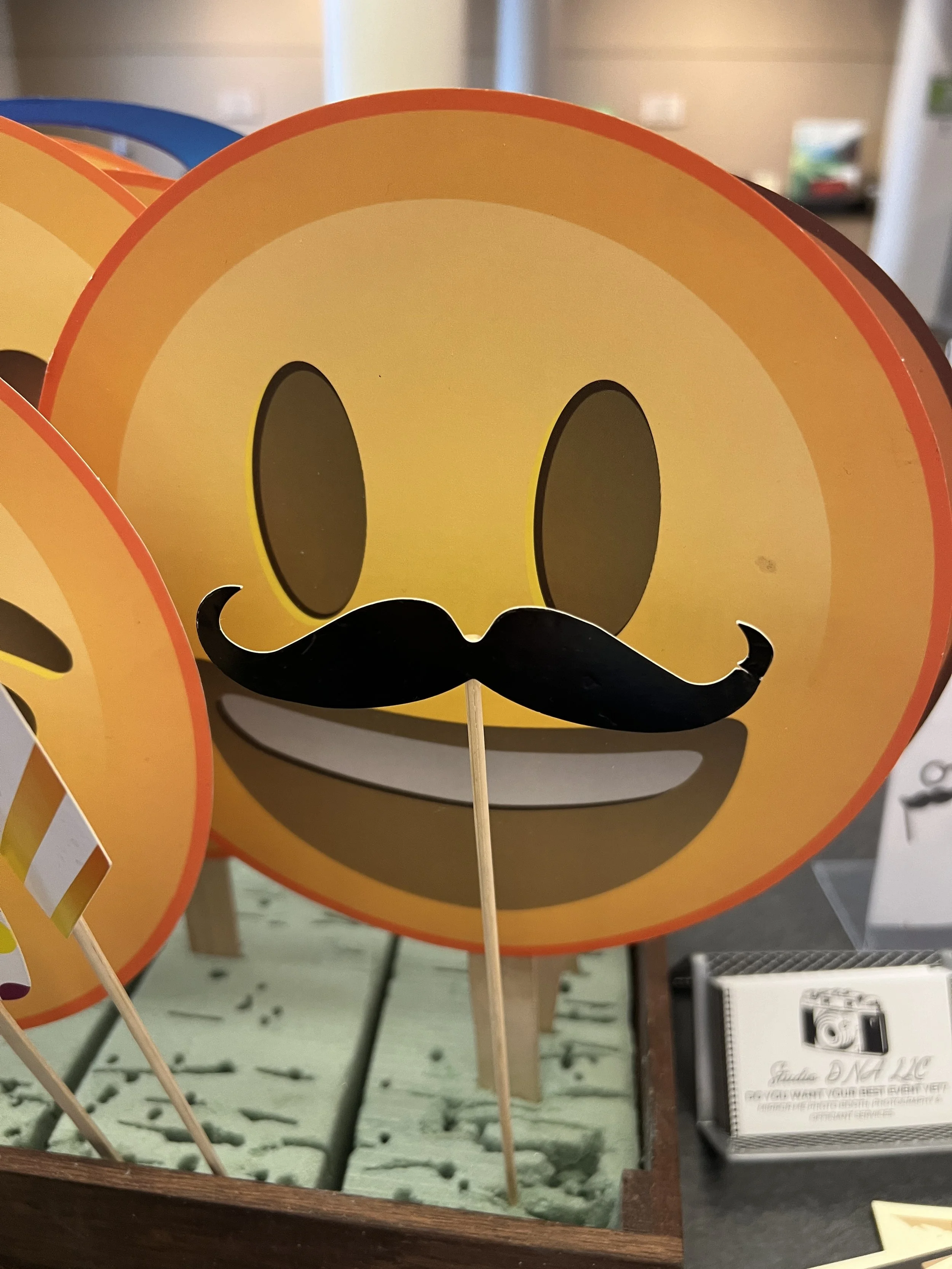 Close-up of a paper smiley face with a moustache, mounted on a stick, displayed in a wooden box with other similar items.