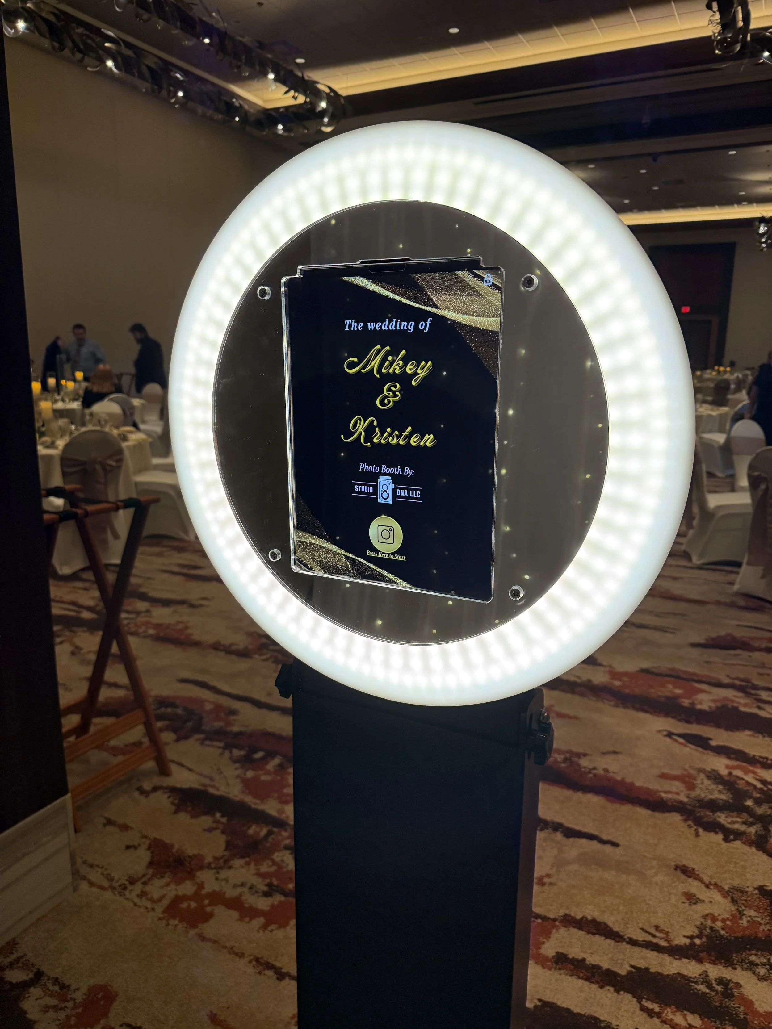 Photo booth with a large illuminated circular frame, displaying a sign that reads 'The wedding of Mikey & Kristen,' in a banquet hall with tables and people in the background.