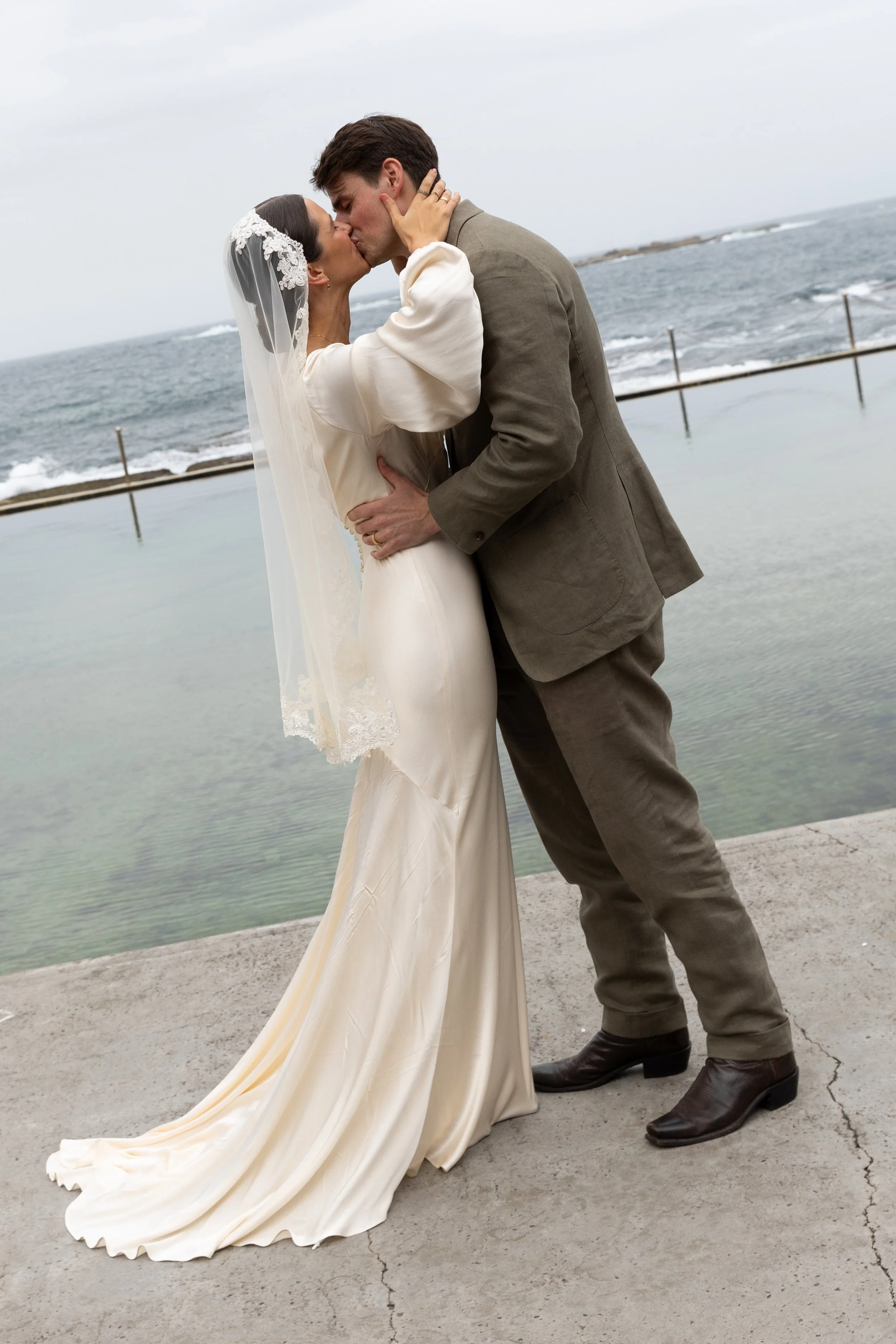 A bride and groom share a kiss by the ocean, with the bride wearing a white wedding dress and veil, and the groom in gray trousers and a brown jacket.