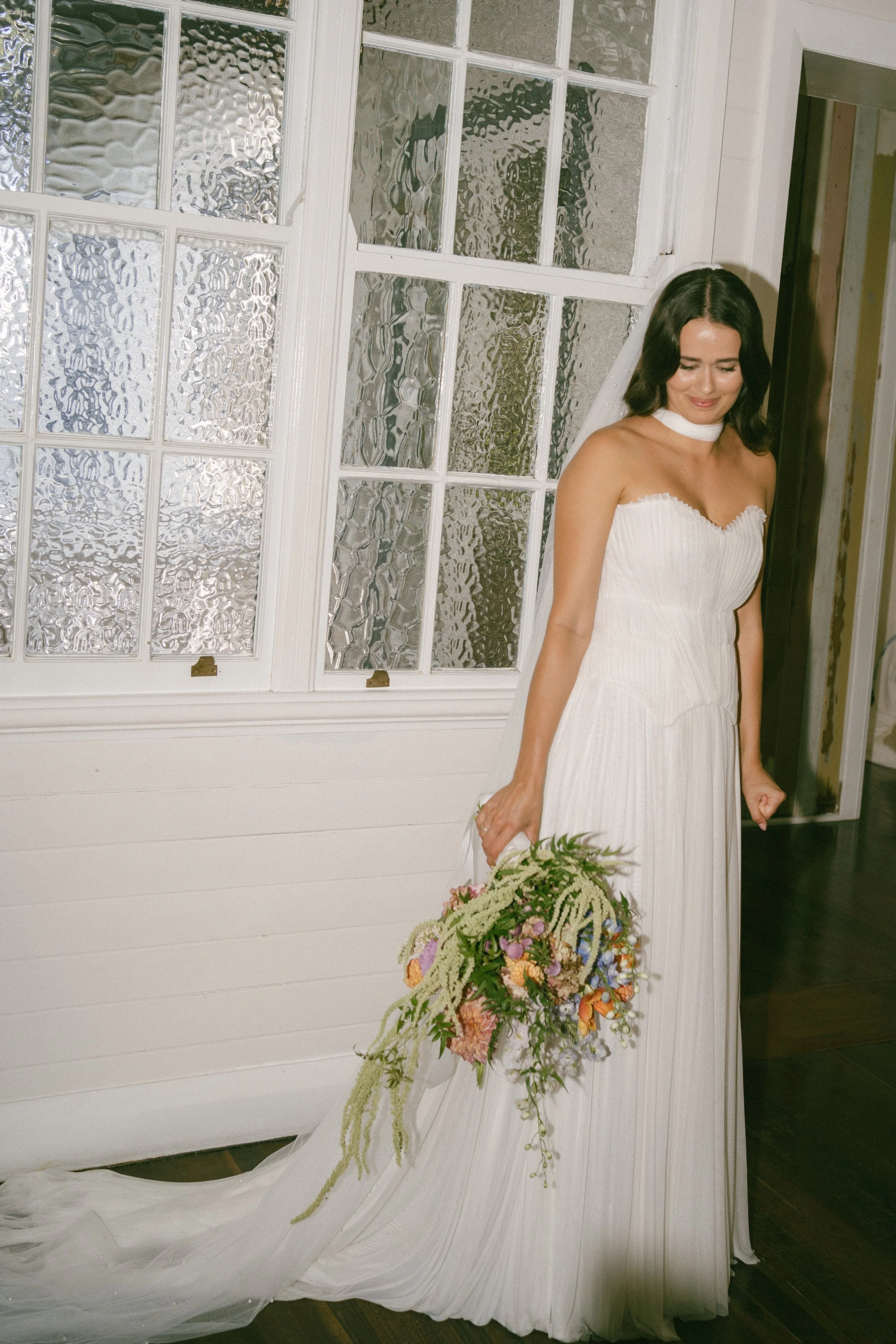 A woman in a white strapless wedding dress with a high slit, holding a bouquet of flowers, stands inside a building near a white-paneled wall and window with textured glass. She appears to be smiling softly with her eyes closed.