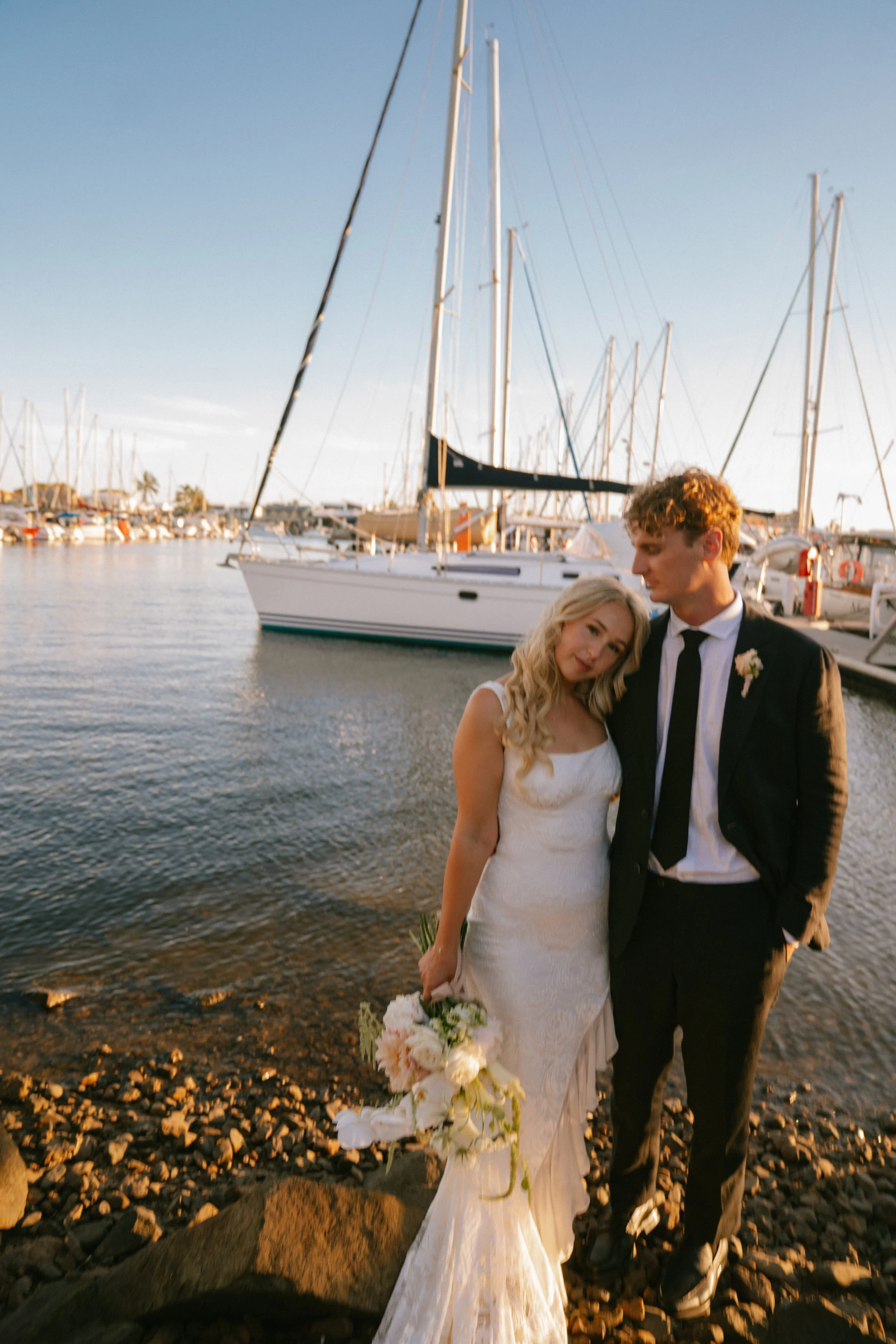 A newlywed couple stands on a rocky shoreline near a marina during sunset. The bride wears a white lace wedding dress and holds a bouquet of flowers, while the groom wears a black suit with a white shirt and black tie. Sailboats are docked in the background.