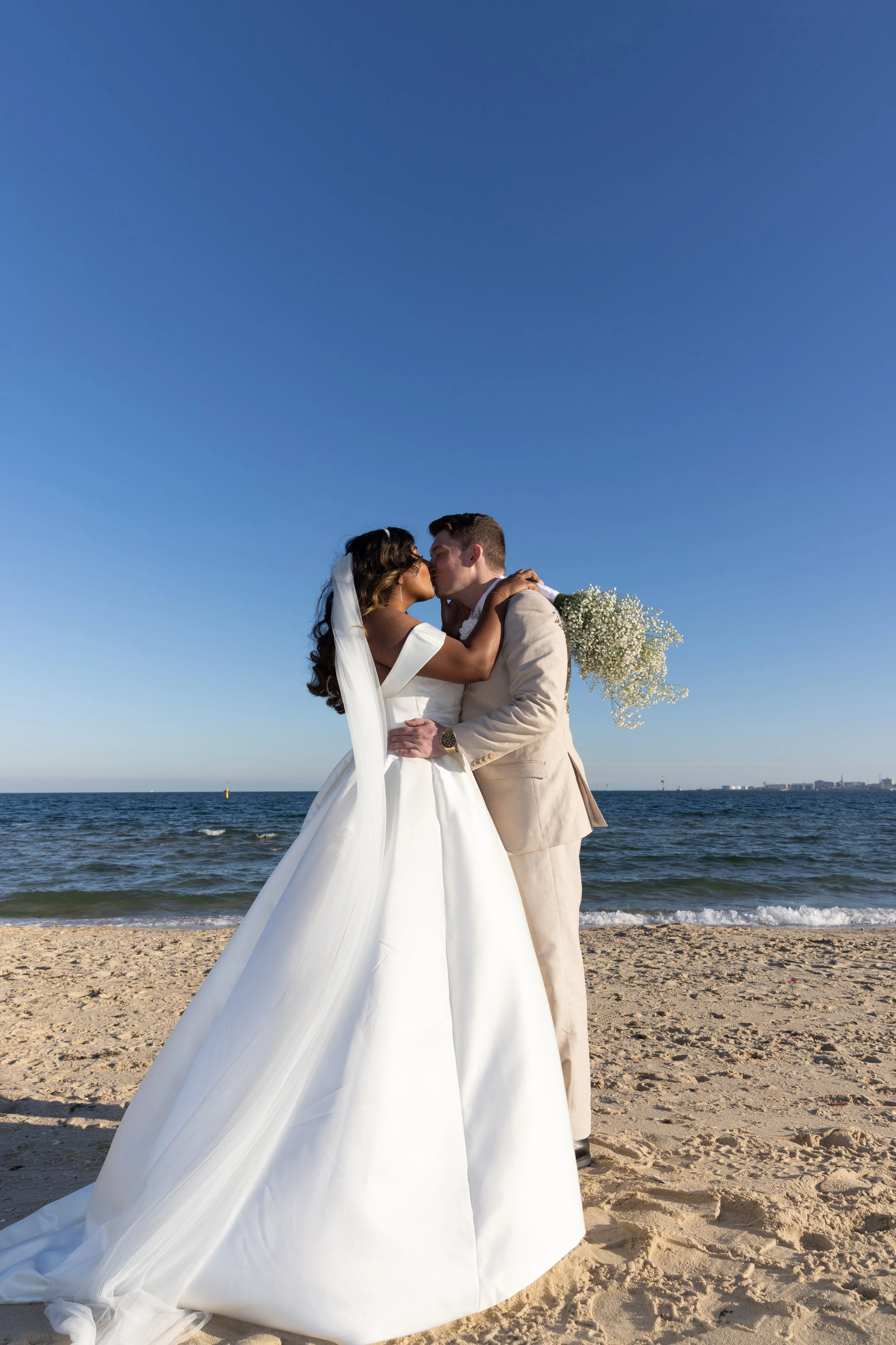 A bride and groom sharing a kiss on the beach, with the ocean and clear blue sky in the background, the bride holding a bouquet of white flowers.