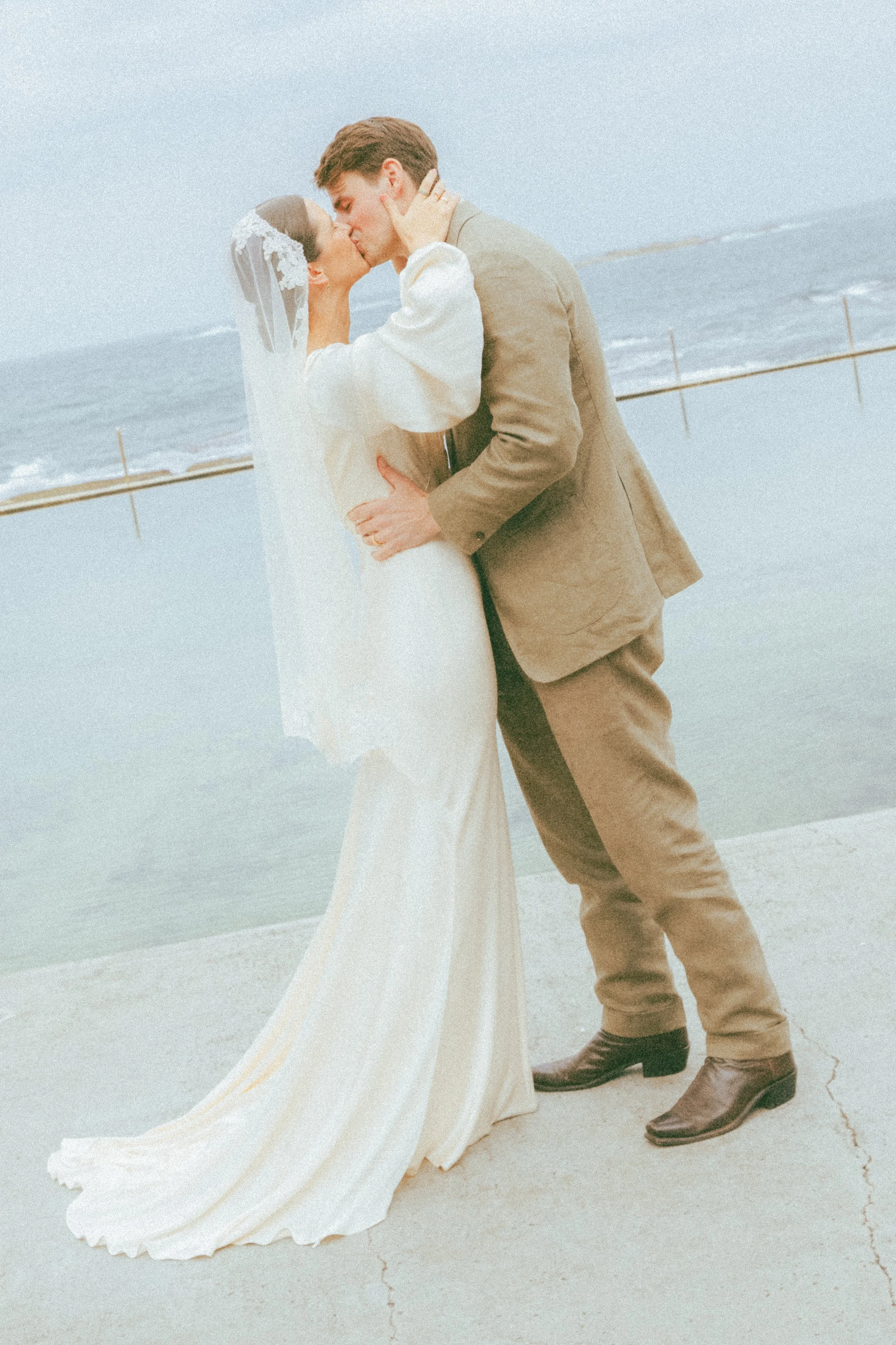 A bride and groom kiss on a beach with the ocean in the background, the bride in a white wedding dress with lace veil, and the groom in a tan suit, brown boots, and black shoes.