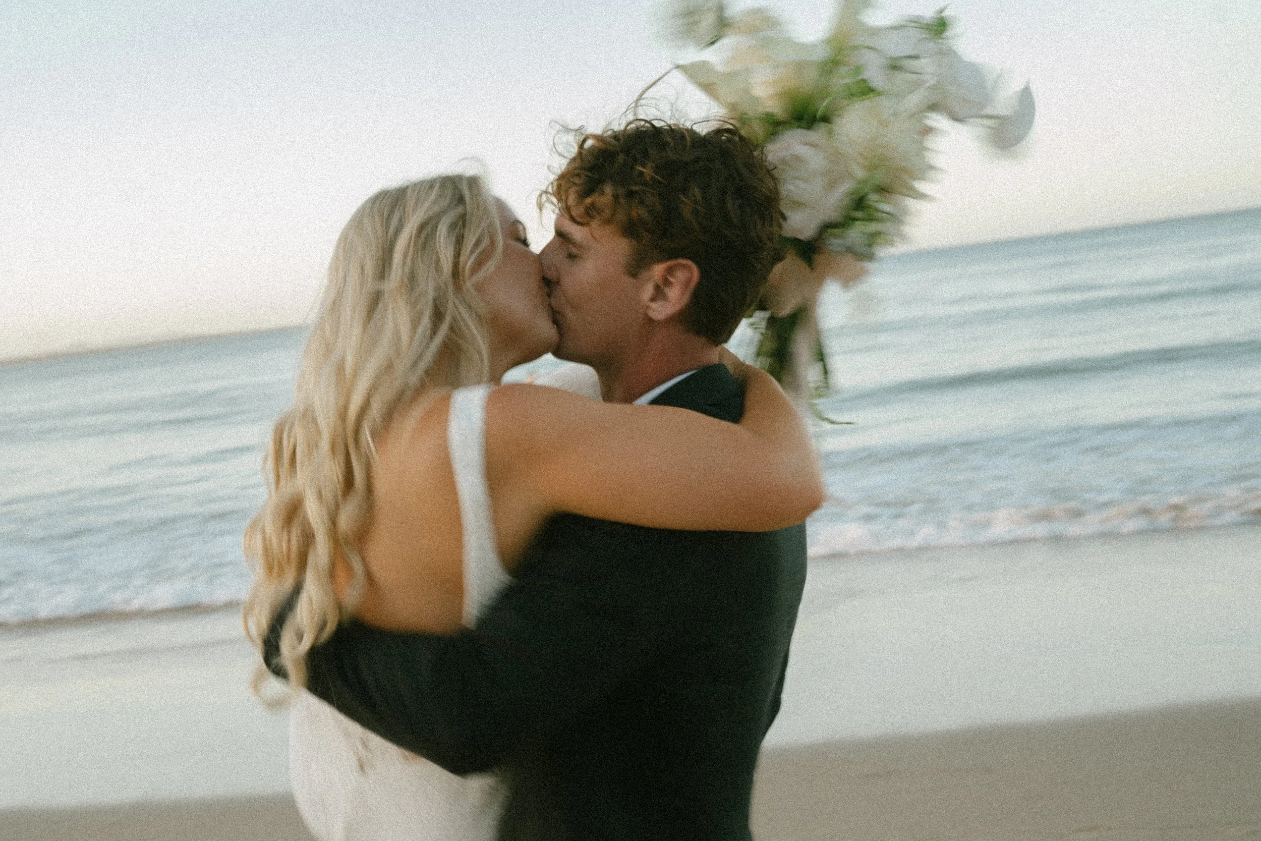A couple kissing on a beach with ocean waves in the background. The woman has long blonde hair and the man has curly brown hair. There is a floral bouquet behind them.