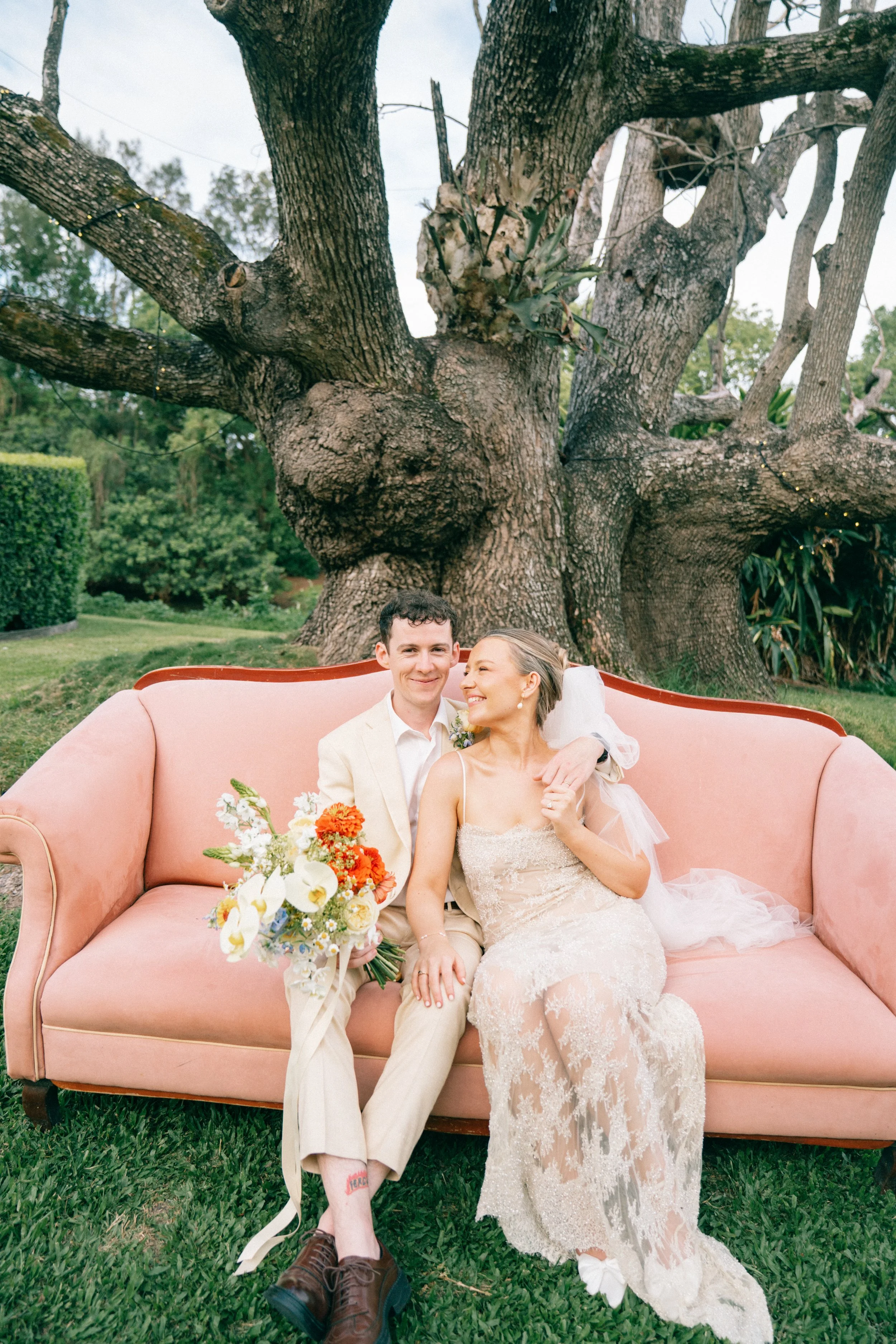 A newlywed couple sitting on a pink vintage sofa outdoors in front of a large tree with green foliage, holding a bouquet of flowers and smiling.
